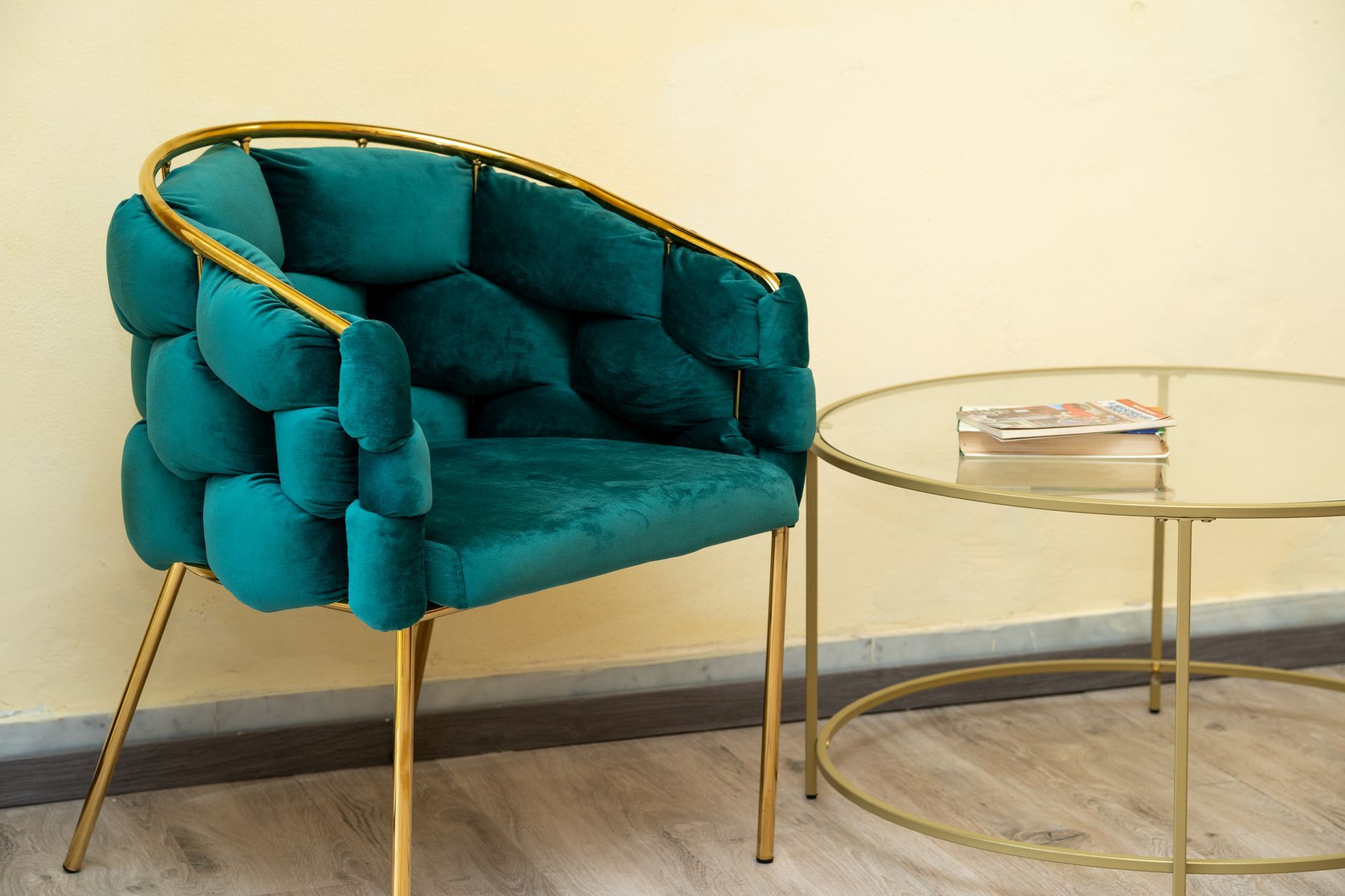 Teal velvet chair with gold frame beside a round glass table with gold legs.