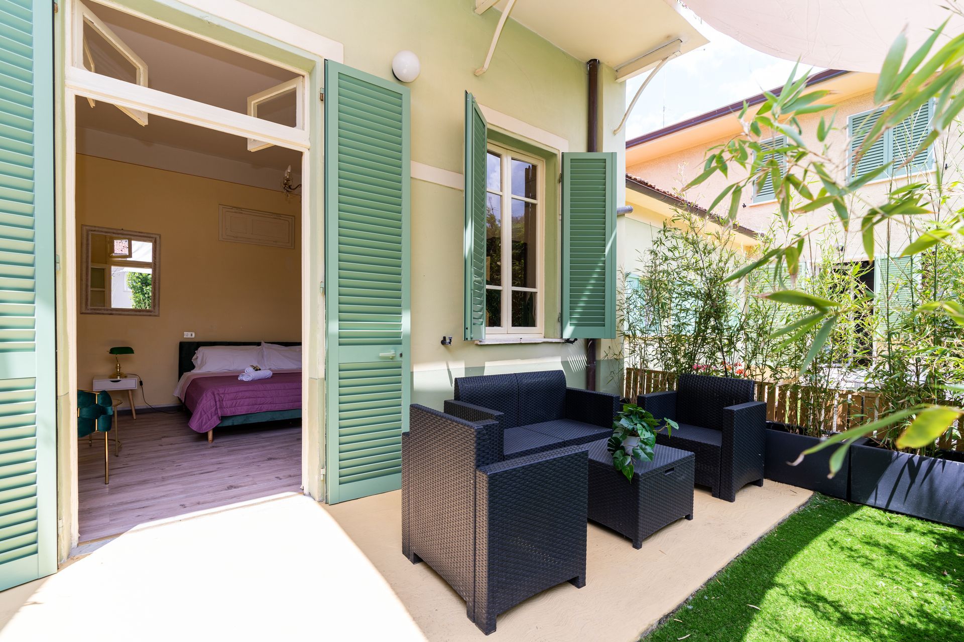 Outdoor patio with black wicker furniture, next to a bedroom with open door and green shutters.