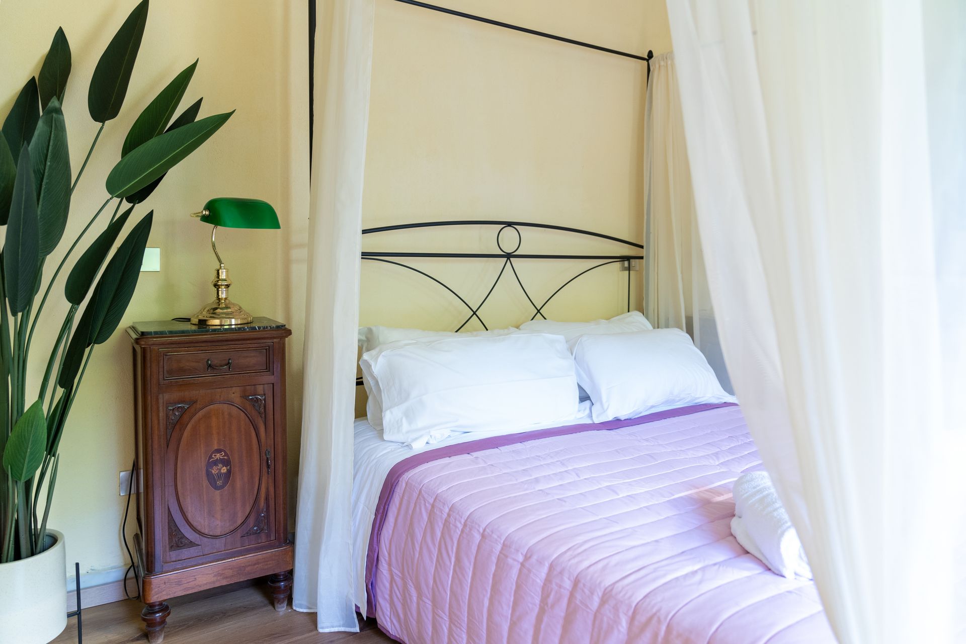 Bedroom with a bed covered in a pink quilt, with sheer curtains, a nightstand, and a potted plant.