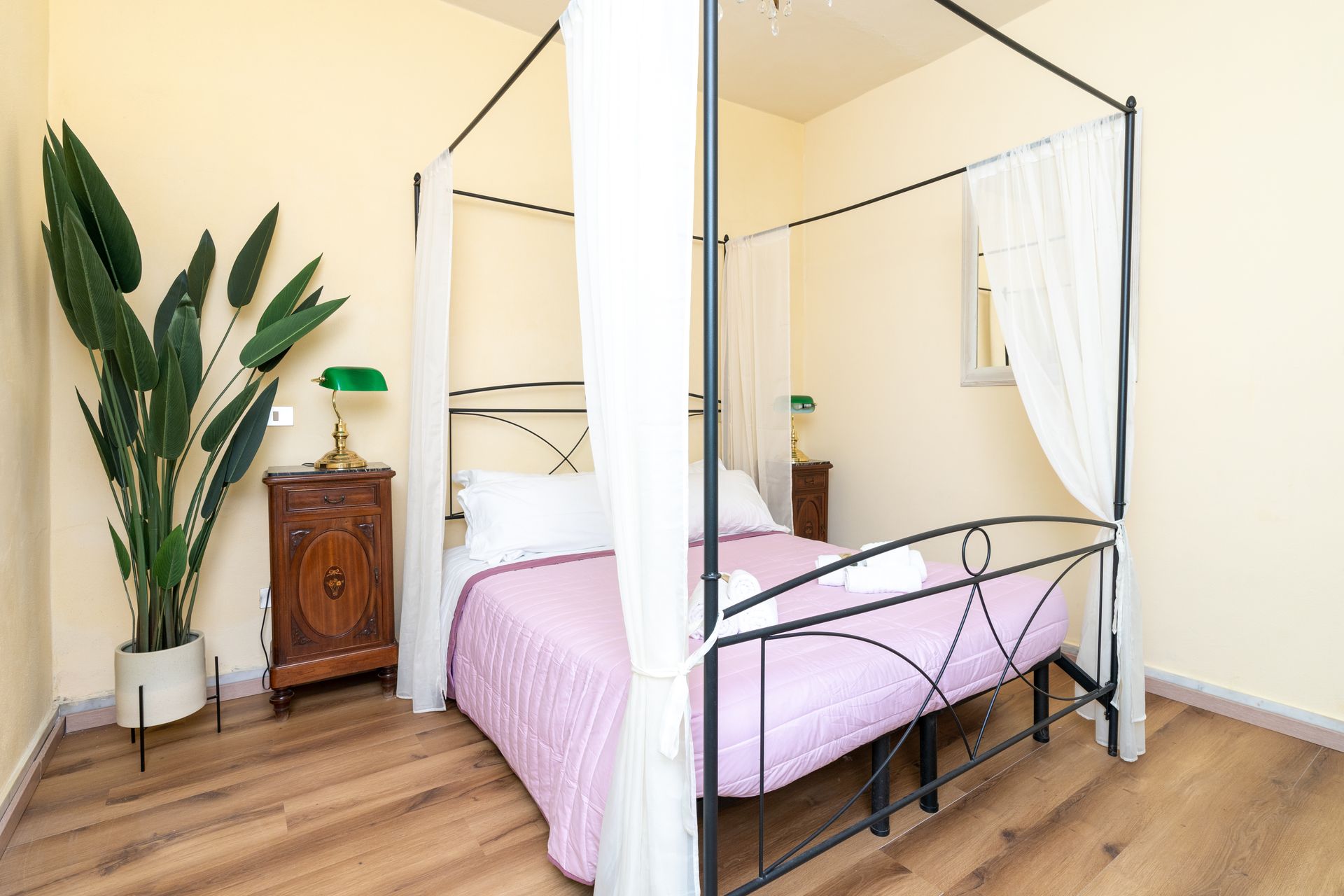 Bedroom with canopy bed, pink quilt, wooden nightstand, green lamp, and large potted plant.