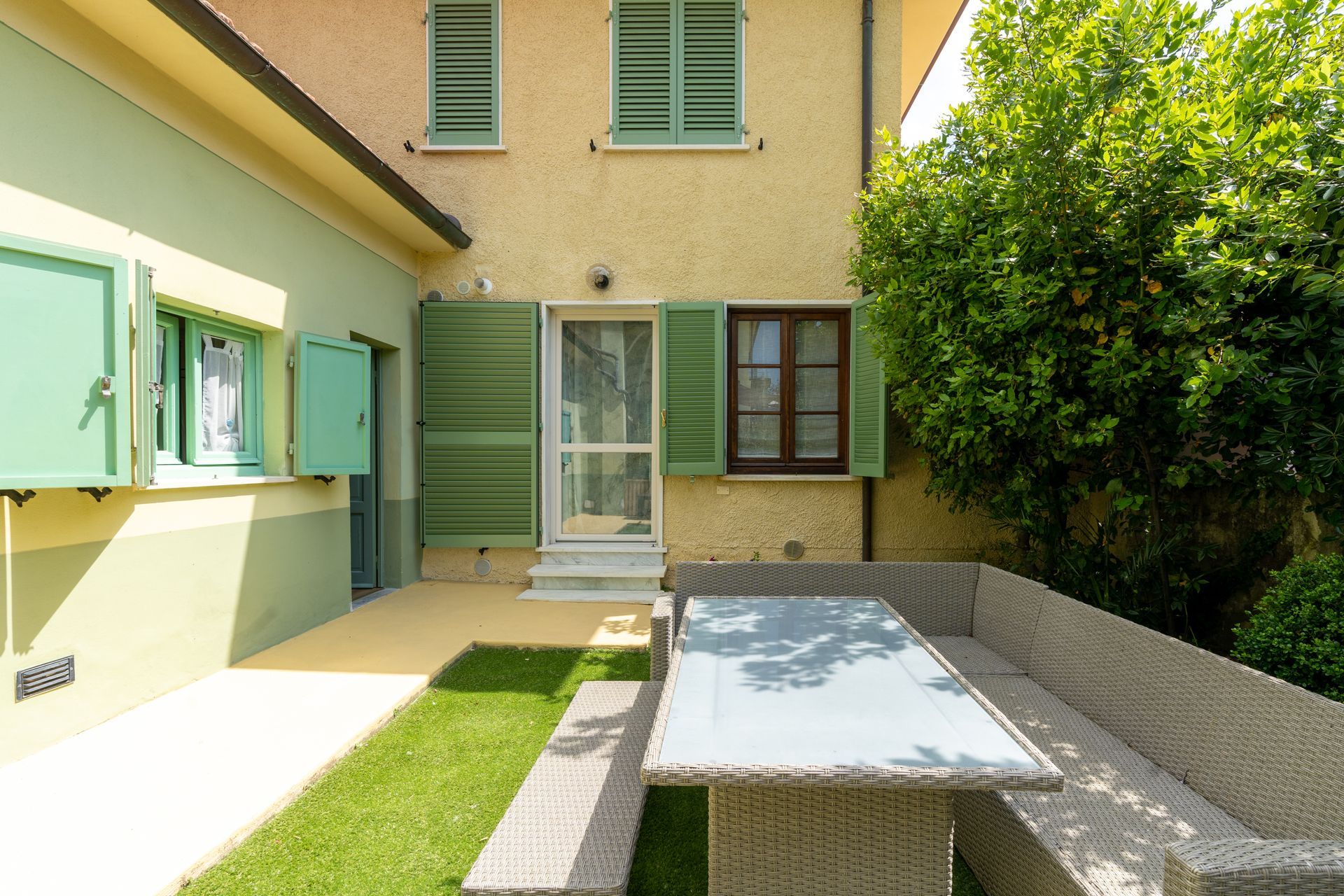 Back patio with a wicker table and benches, next to a building with green shutters.