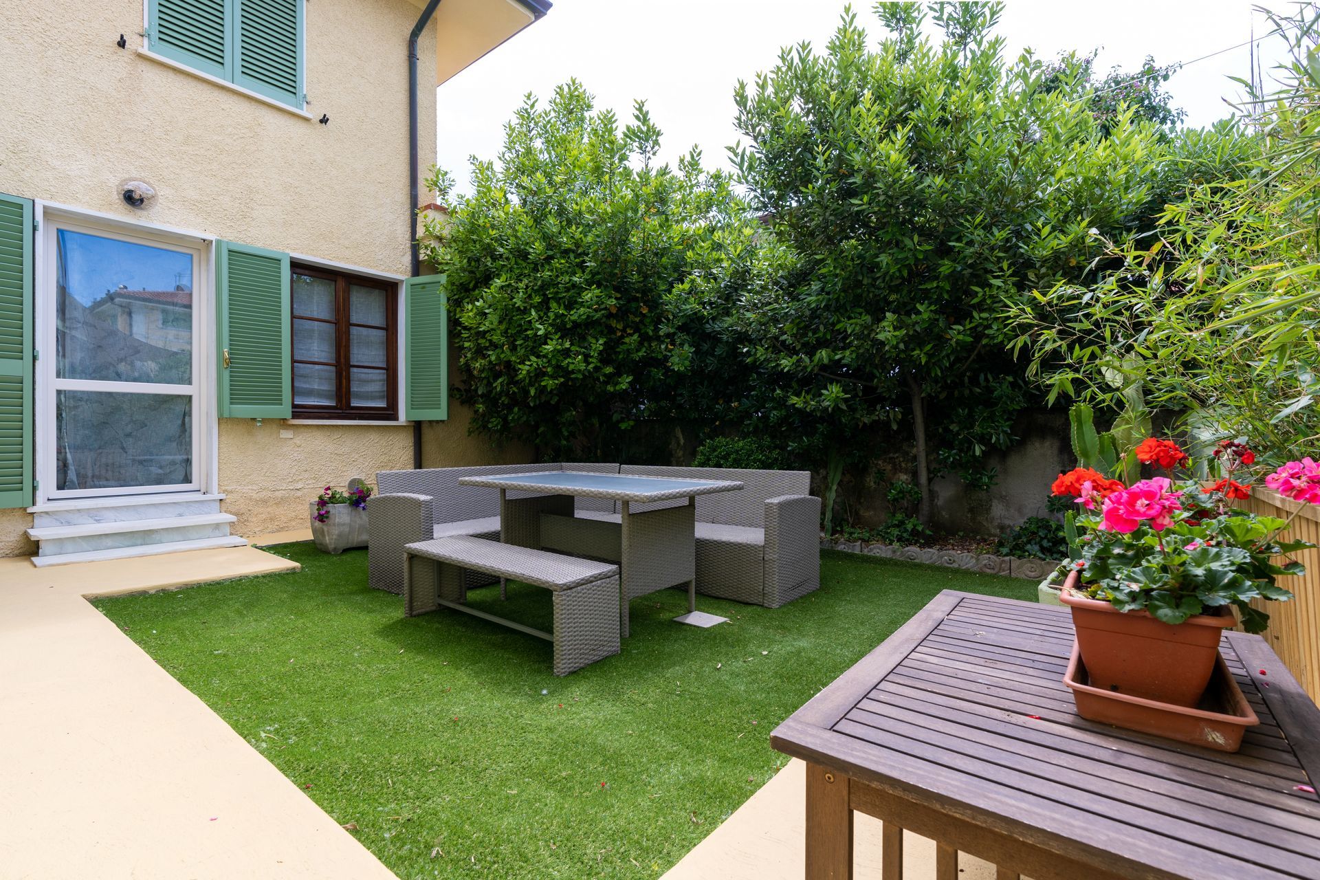 Lawn and patio with outdoor furniture, and flowering plants. House with shuttered windows in background.