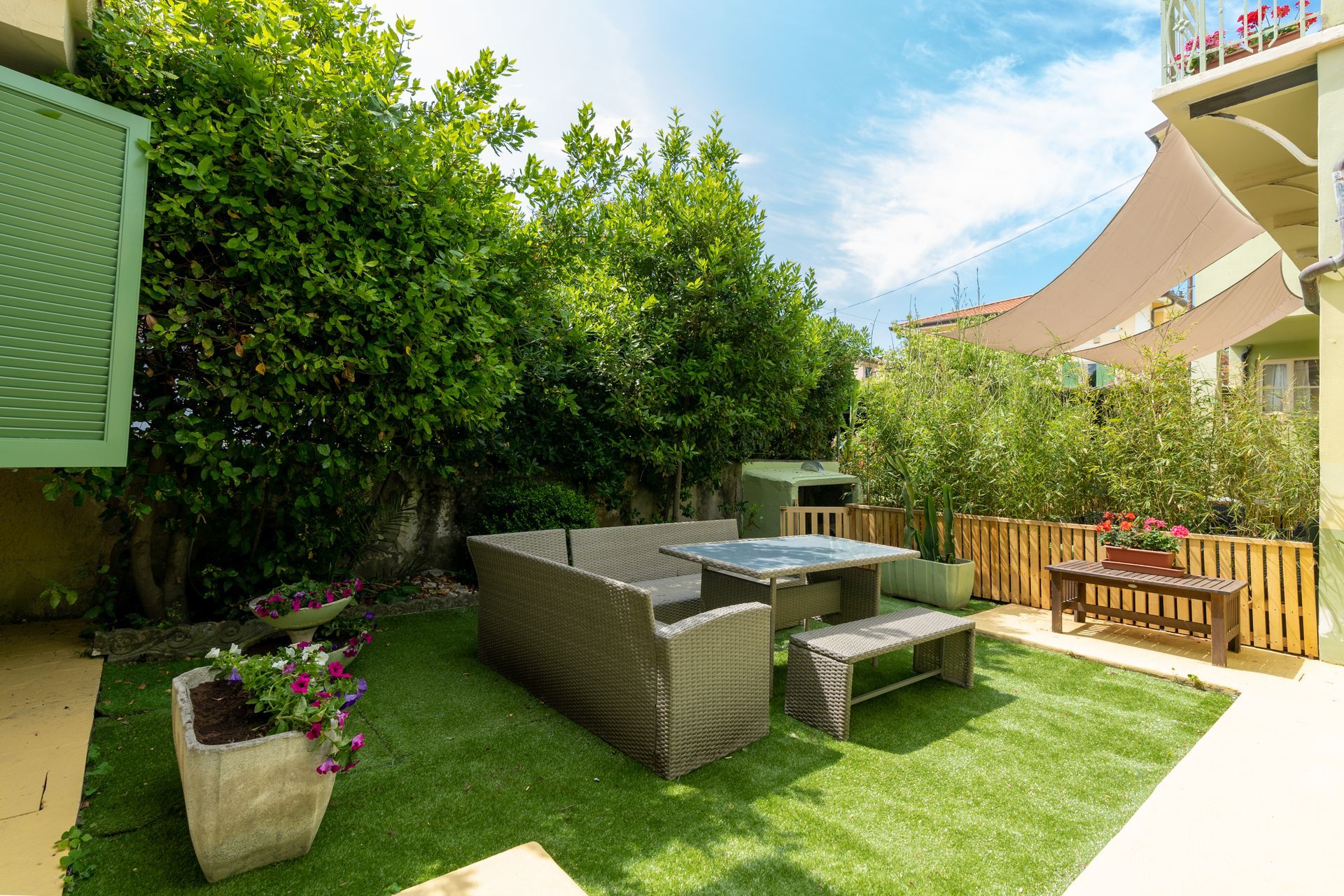 A green, artificial lawn with a wicker patio set. Lush green foliage surrounds the space, with a sunny sky overhead.