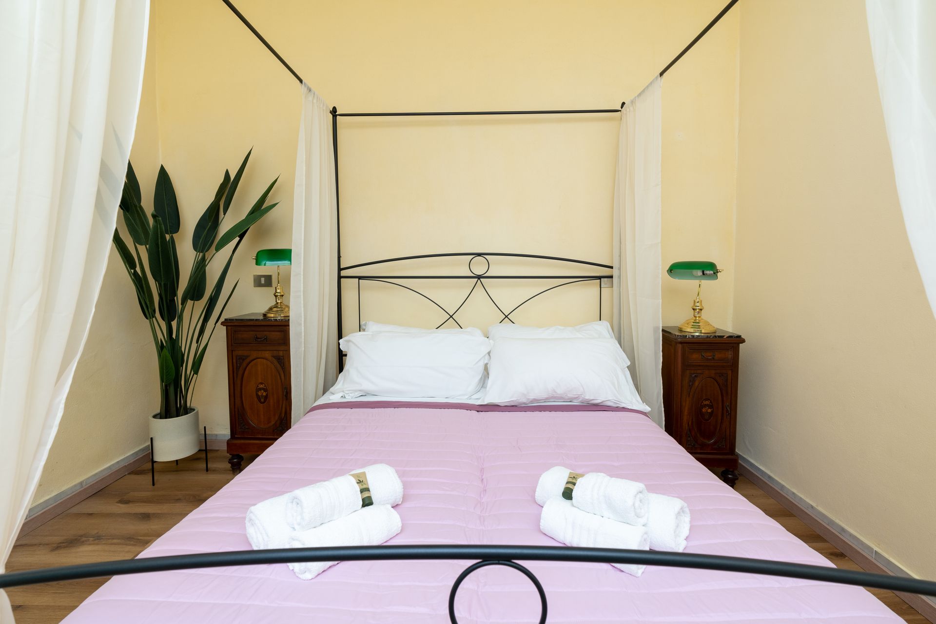Bedroom with four-poster bed; lavender sheet, white pillows and towels, wooden bedside tables, potted plant.