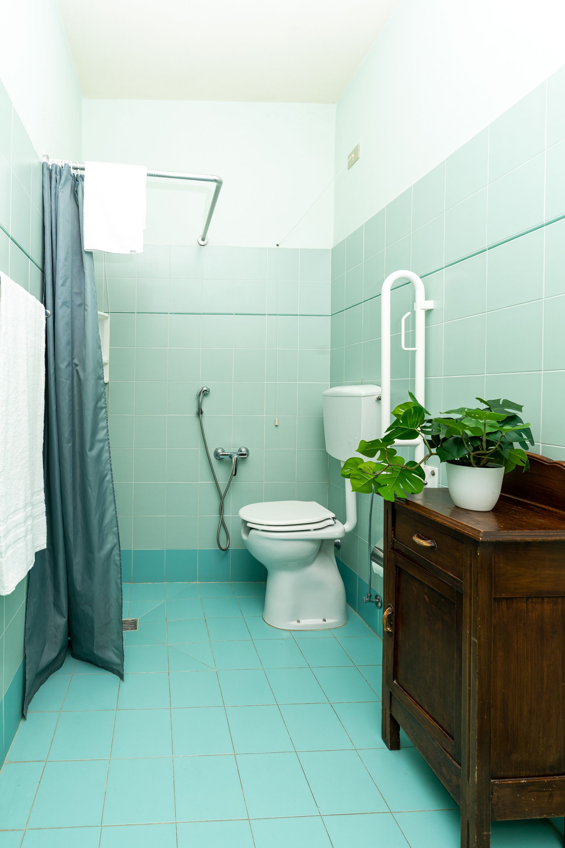 Small bathroom with turquoise tile walls and floor. Toilet, shower curtain, sink with plant, and grab bar.