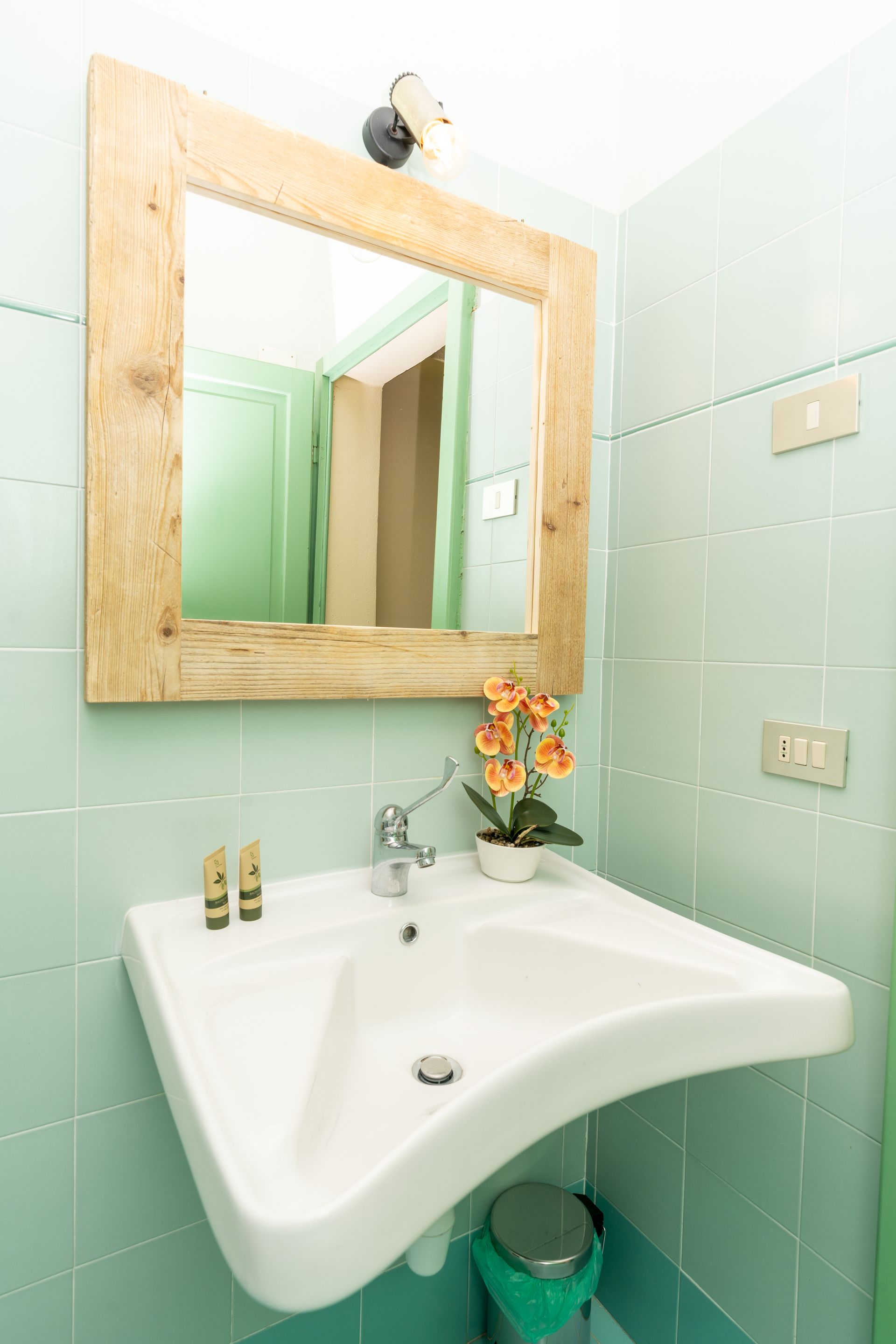 Bathroom with a square wood-framed mirror above a white sink, featuring a small orange flower and light blue tiles.