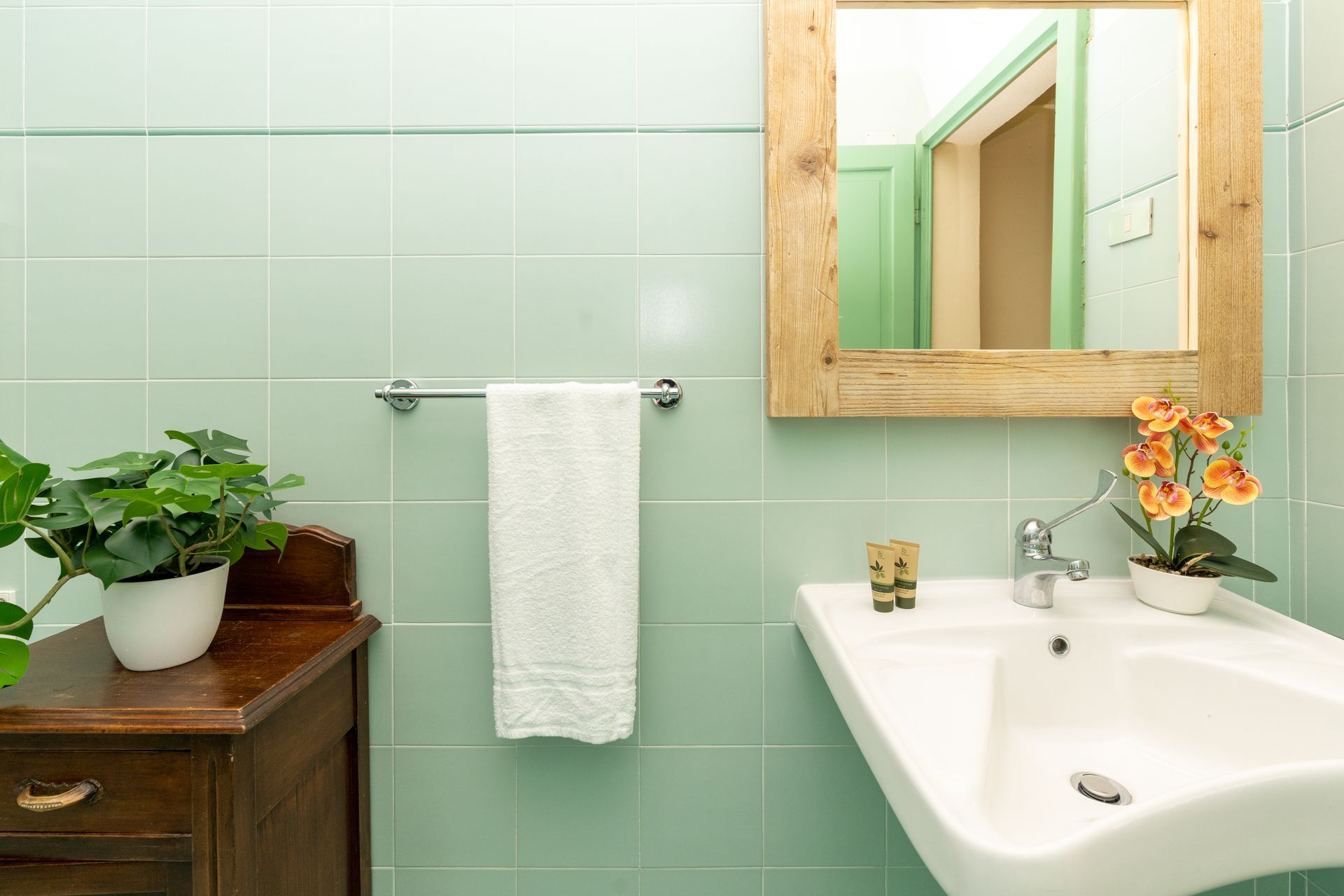 Green-tiled bathroom with a wooden cabinet, towel rack, sink, mirror, and plants.