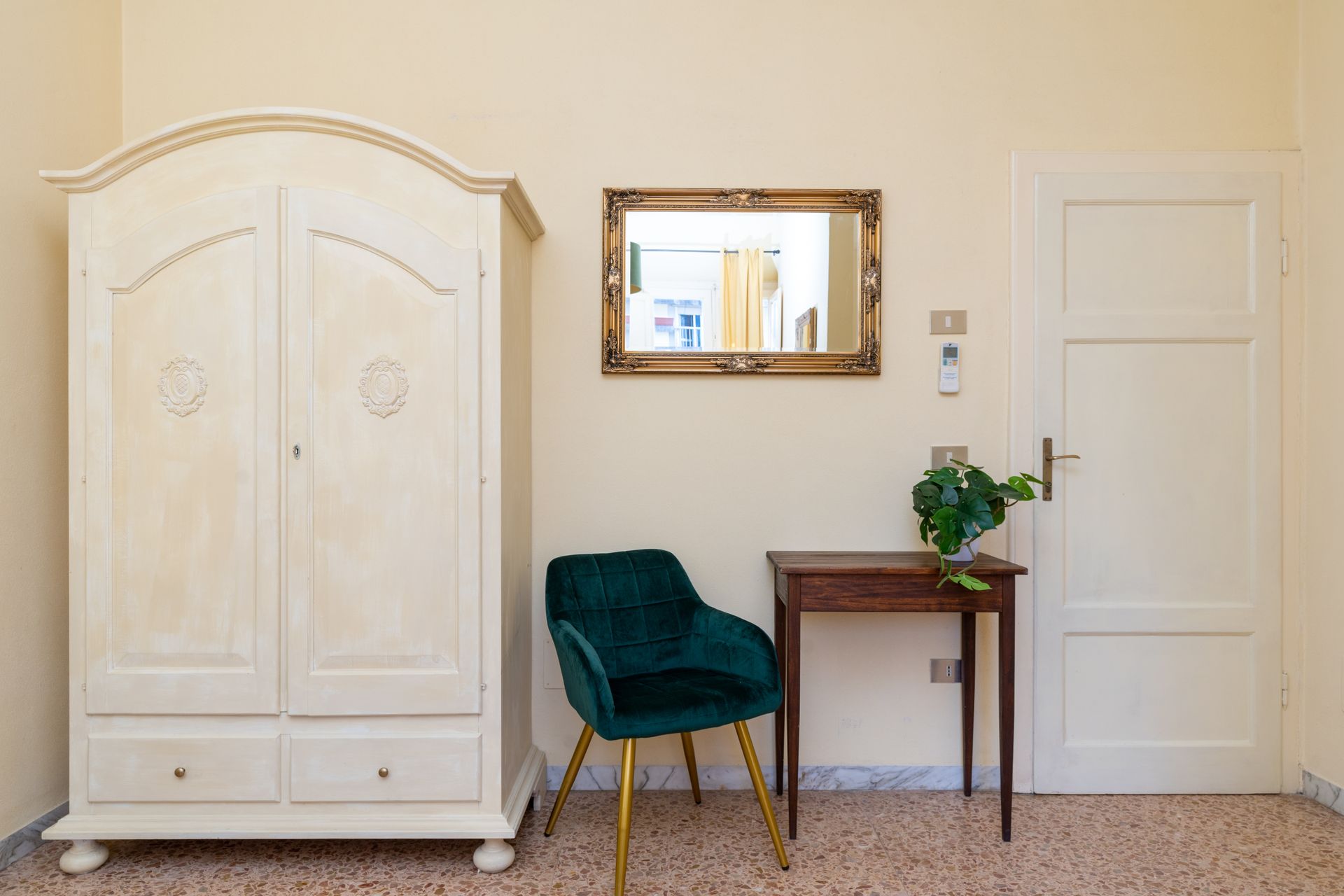 Cream wardrobe, ornate gold-framed mirror, green velvet chair, small wooden table with plant, and white door in room.