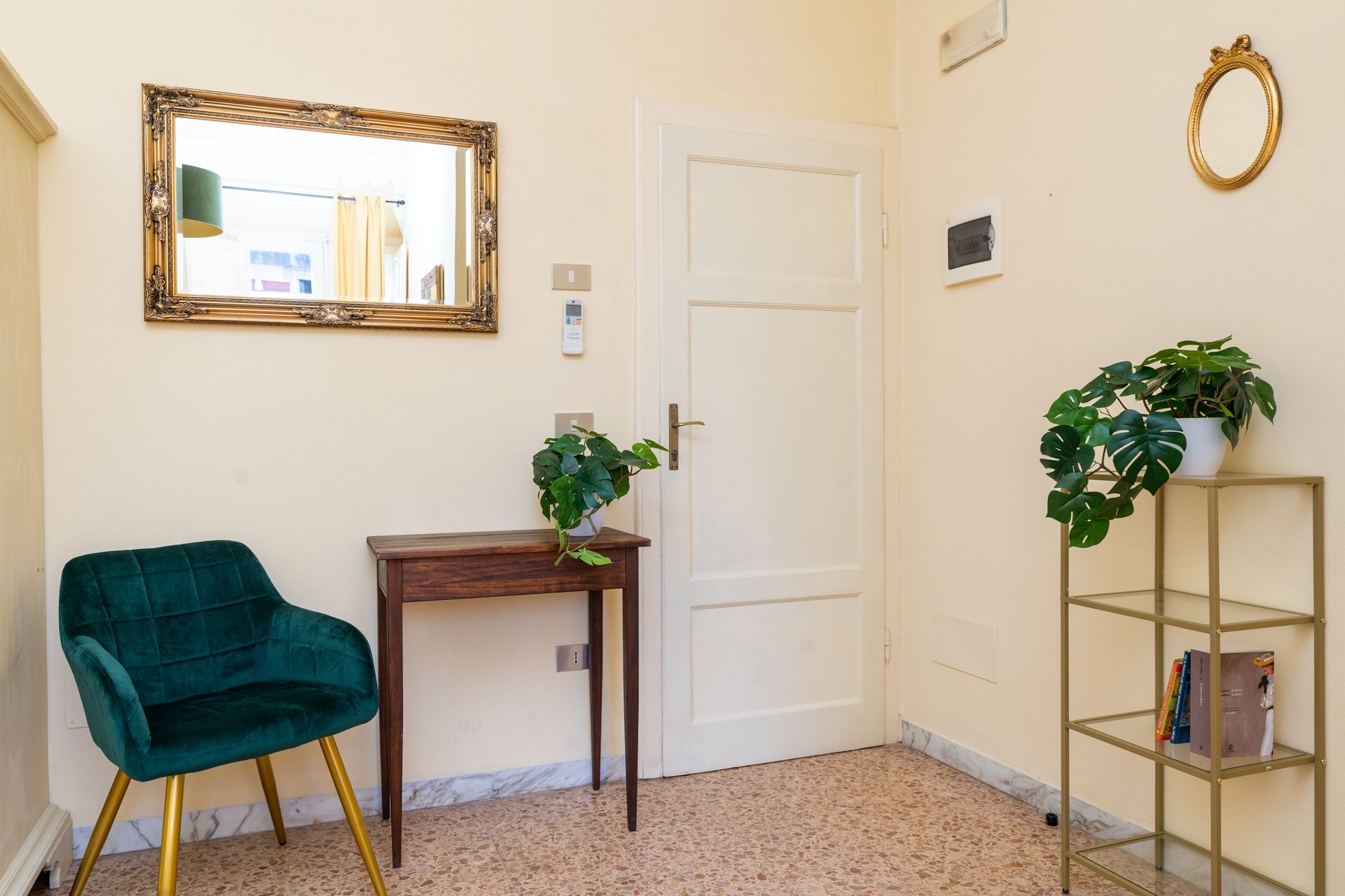 Hallway with green chair, small table, gold shelves, and mirror. White door and wall with plants.