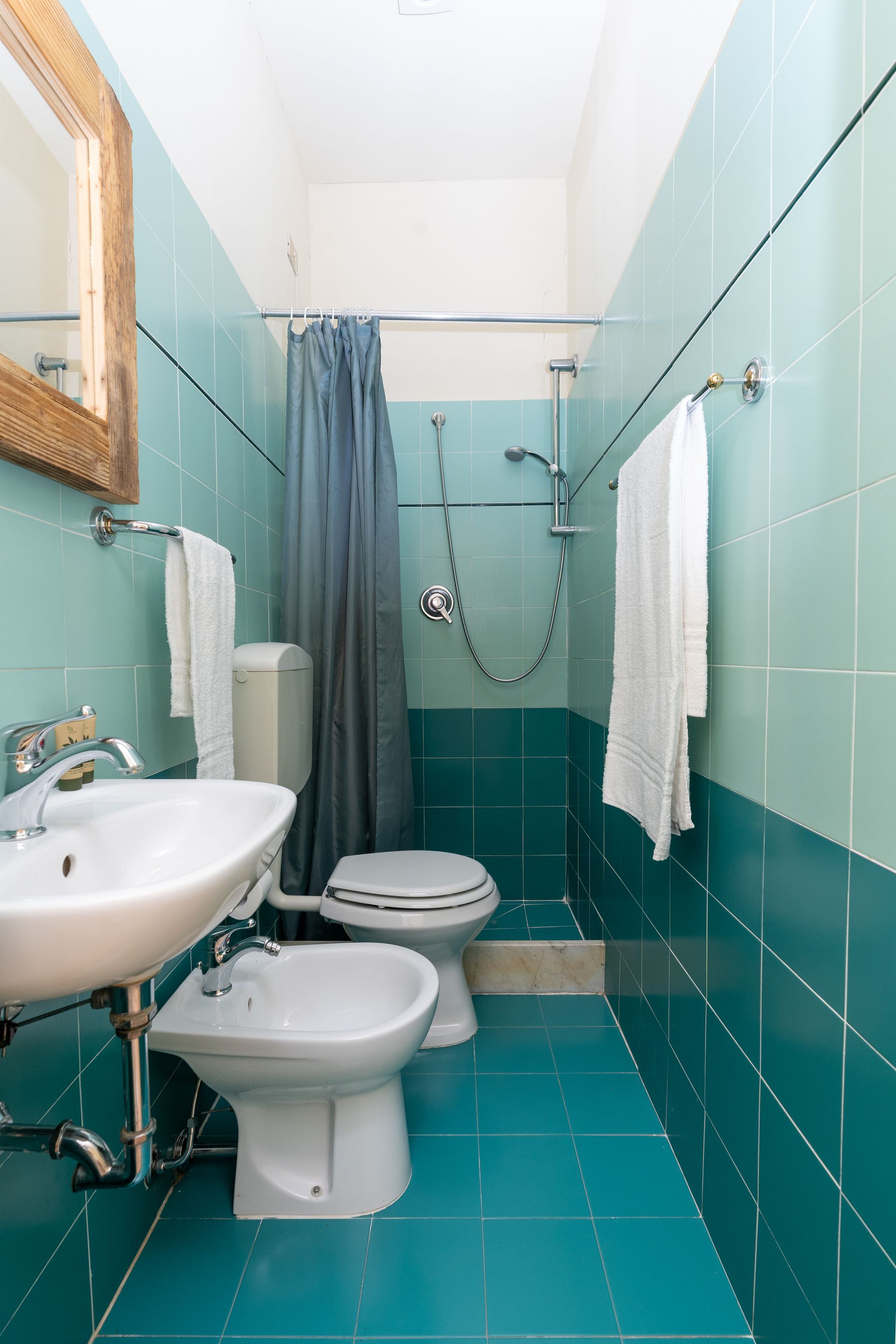 A narrow bathroom with turquoise tile. Sink, toilet, bidet, shower, and towels are visible.