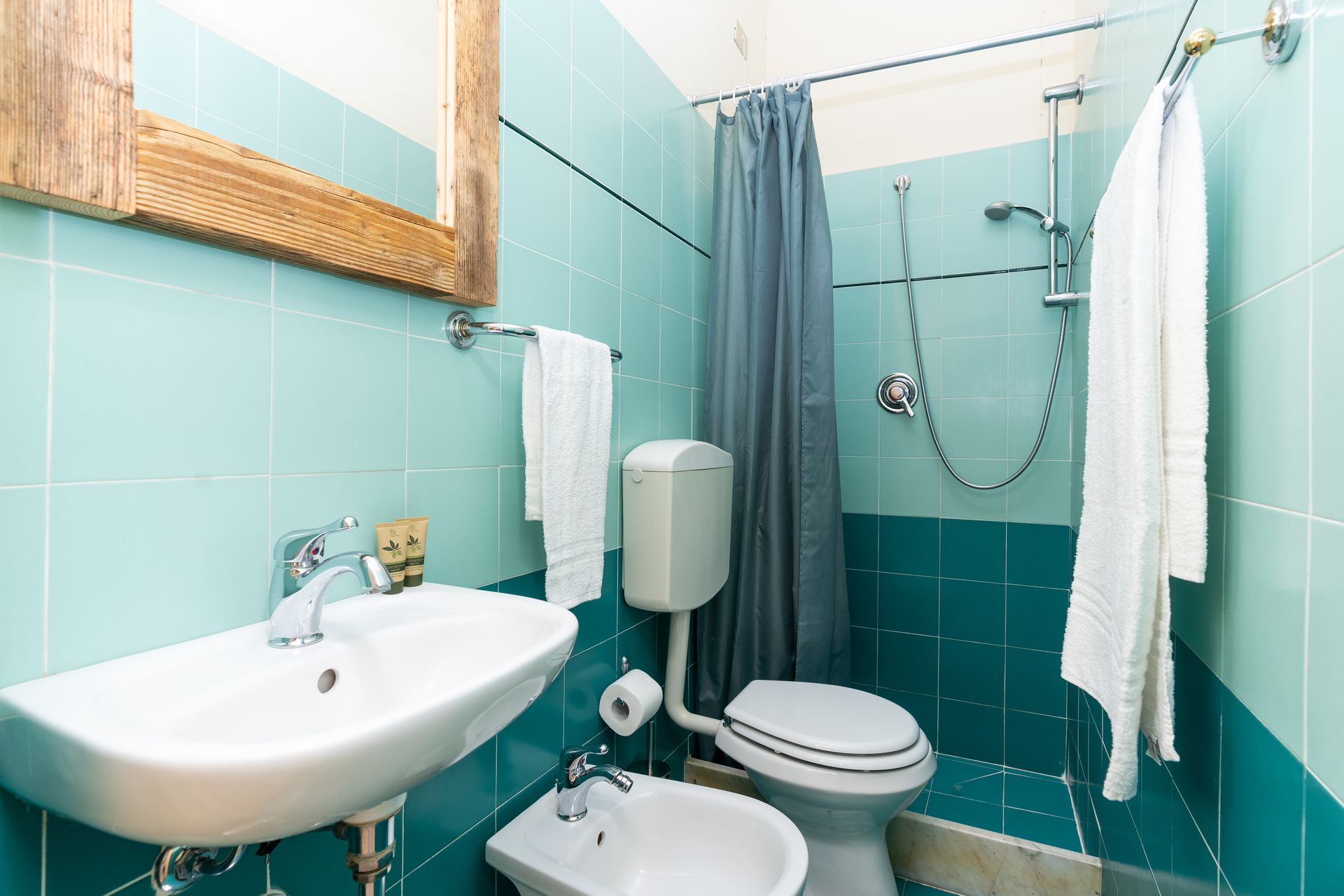 Bathroom with turquoise tile, white sink and toilet, shower with blue curtain, gold-framed mirror, and white towels.