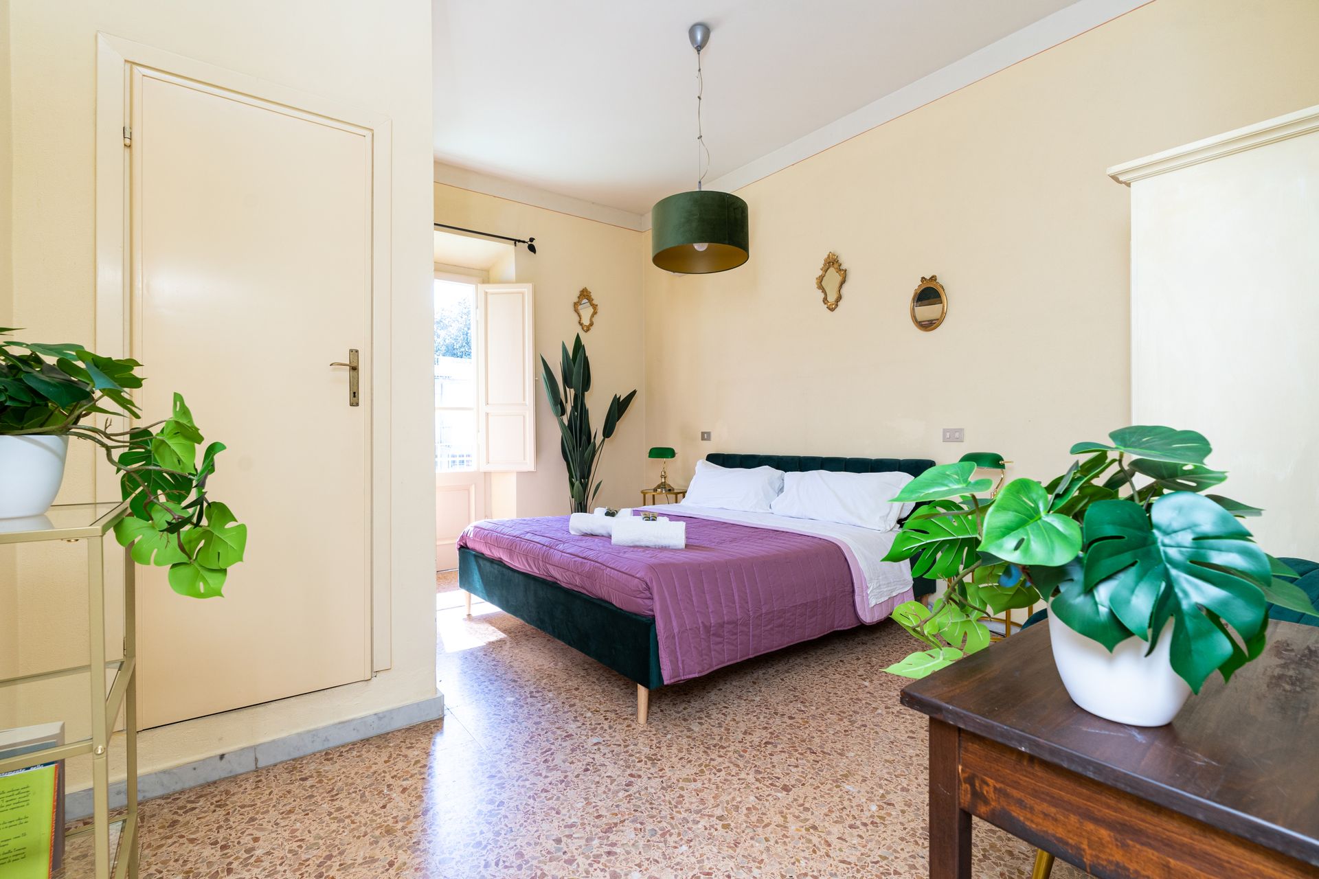 Bedroom with bed, plants, and natural light coming through a window. Cream walls and patterned floor.