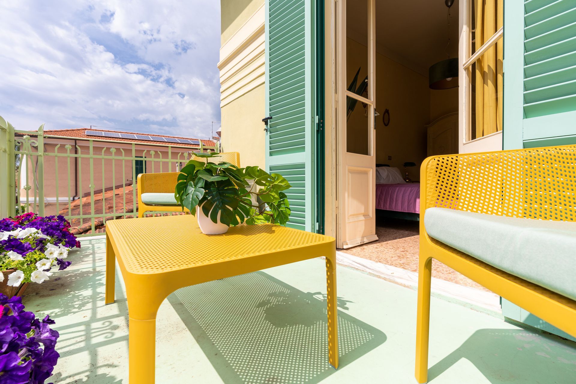 Balcony with yellow furniture, a potted plant, and purple flowers; door leads to a bedroom.