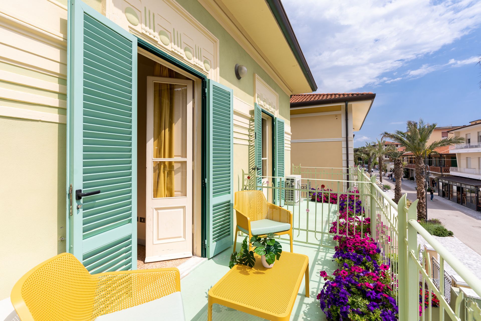 Balcony with yellow furniture, green shutters, and a street view with palm trees.
