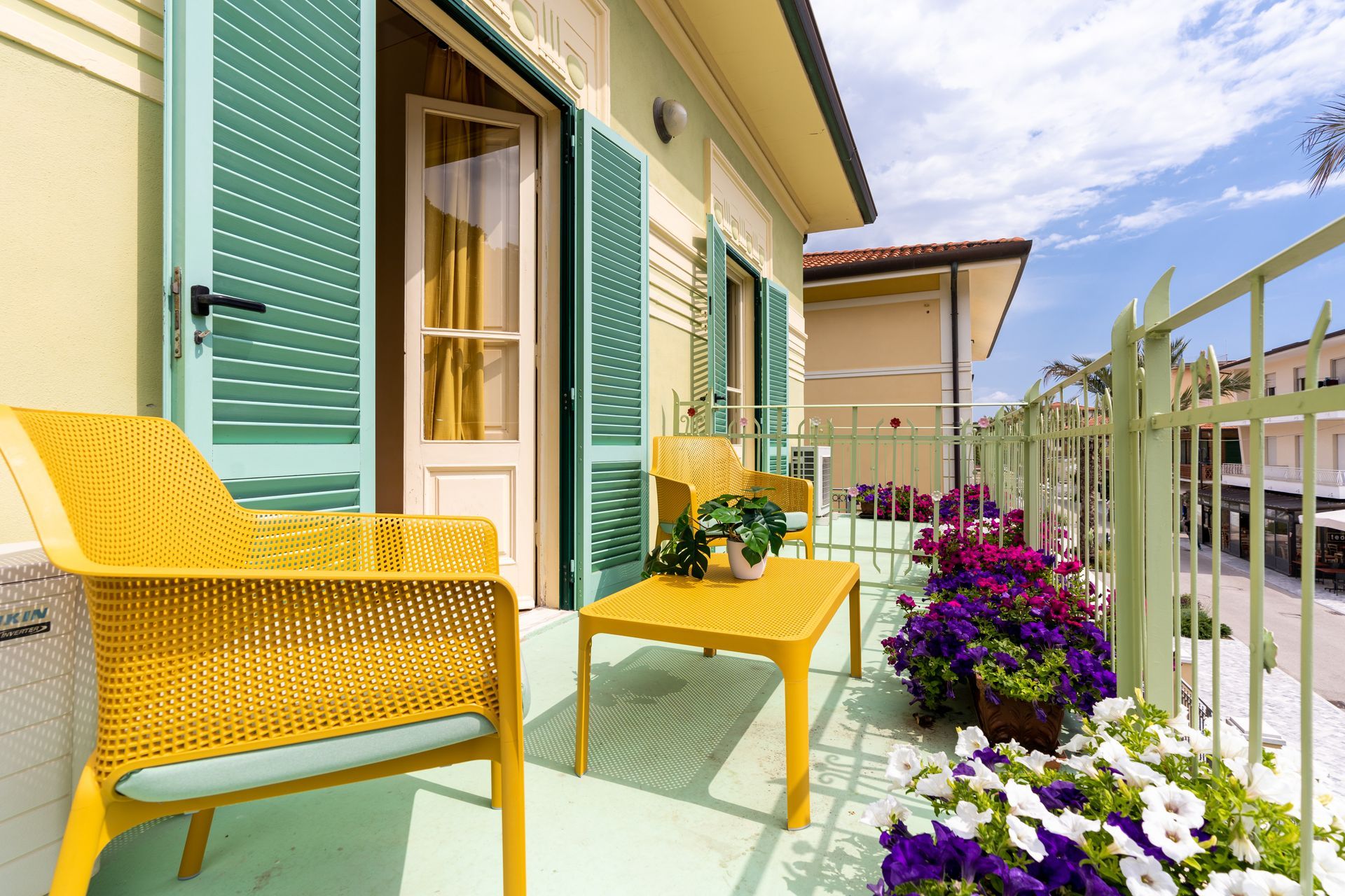 Balcony with yellow furniture, green shutters, and flower boxes; blue sky.