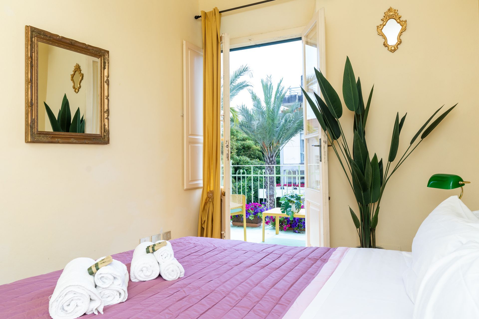Bedroom with open balcony doors, bed with purple cover, gold mirrors, and a large plant.