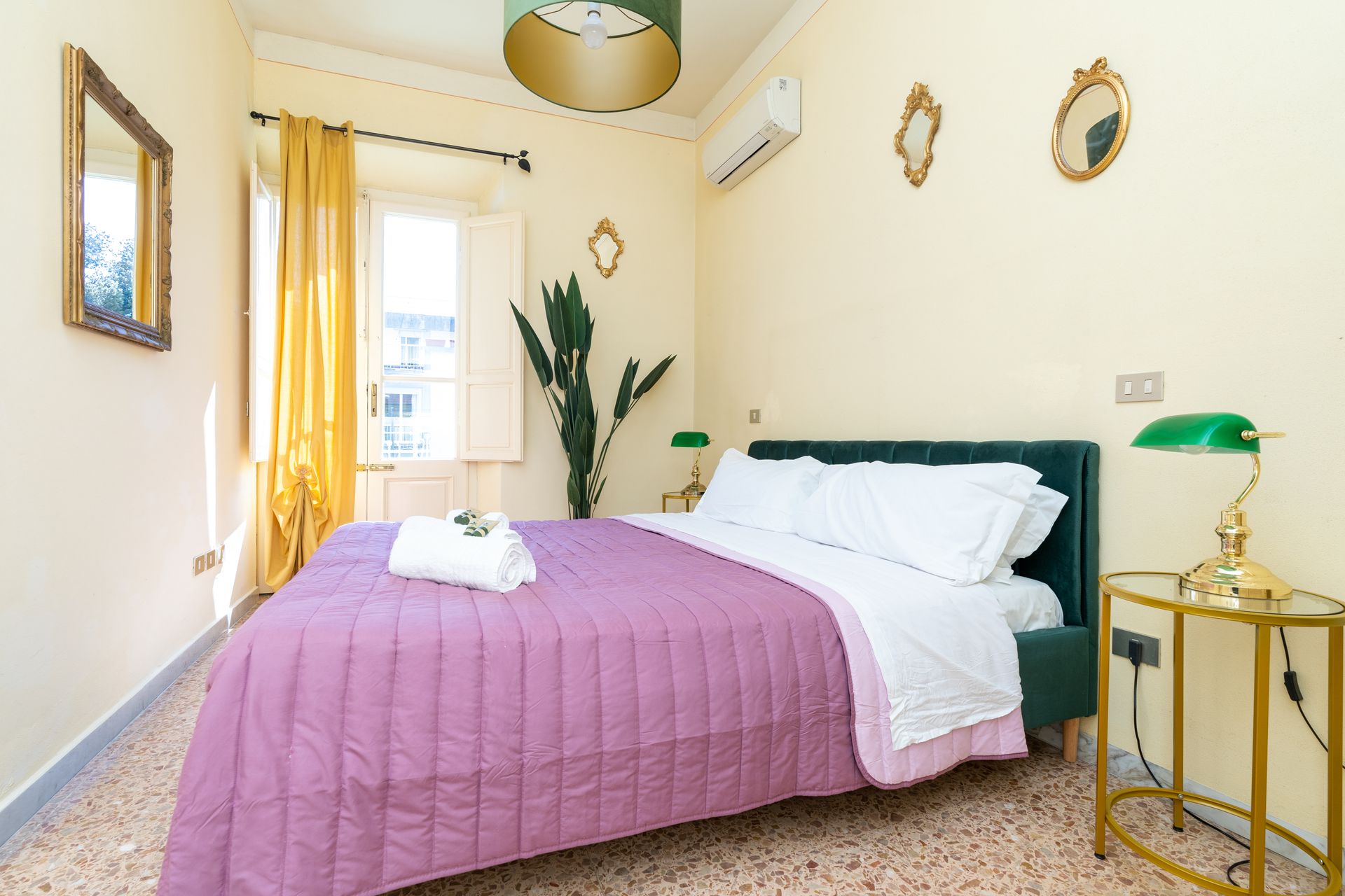 Bedroom with a bed, end table, window, and gold accents. Mauve bedspread, green lamp, and mirrors.
