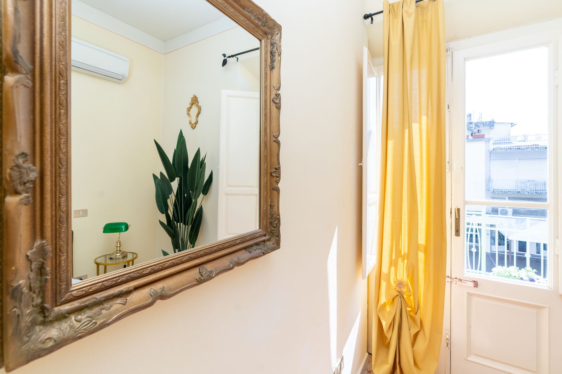 Room with large mirror, yellow curtains, and a door to a balcony. A potted plant and ornate side table are also visible.