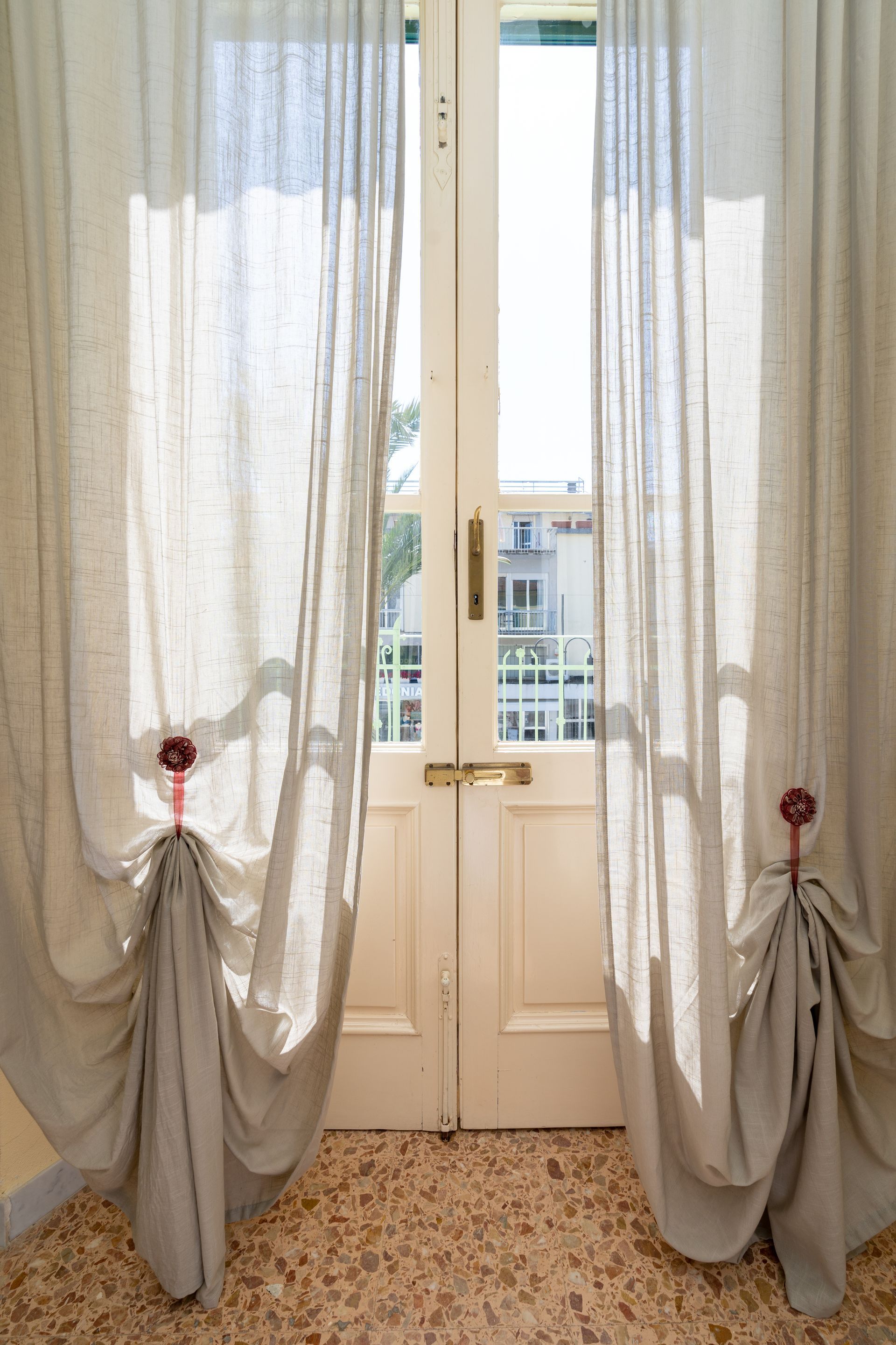 Cream-colored curtains framing an open white door, sunlight streams in, revealing a cityscape.