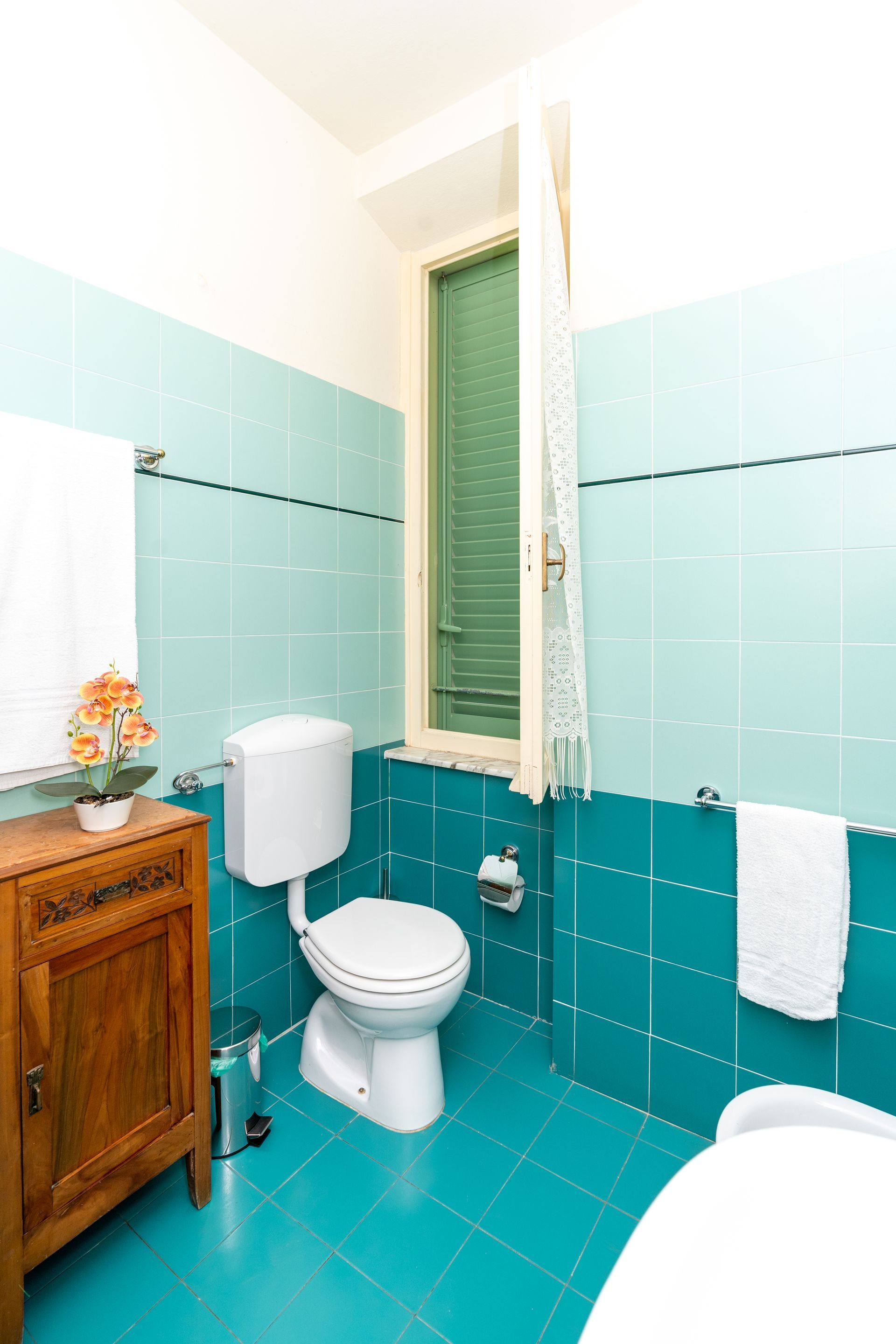 Teal tiled bathroom with toilet, wooden cabinet, and window with green shutters.