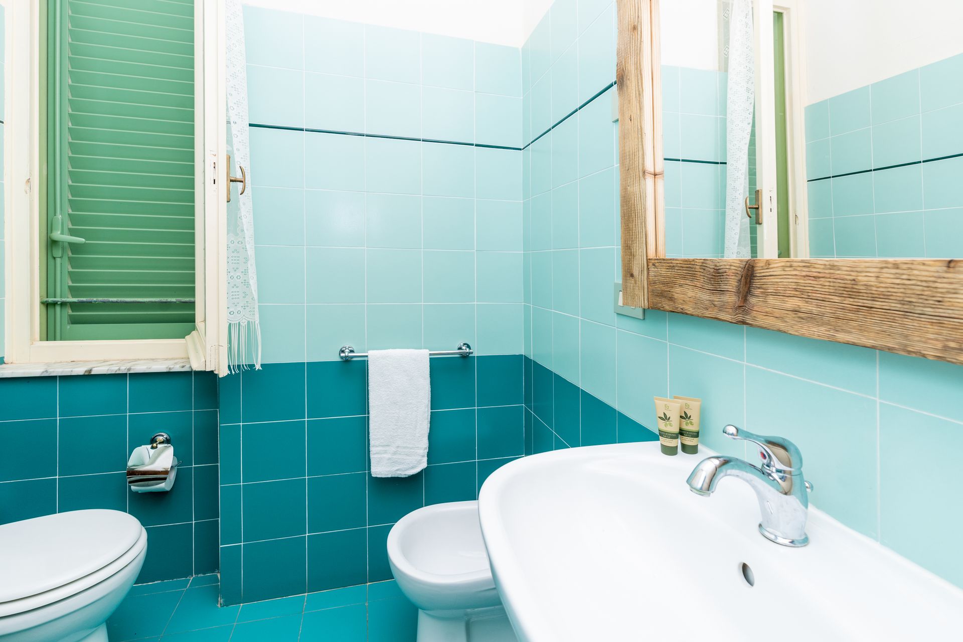 Bathroom with turquoise tiled walls, white sink, toilet, and a window. A towel hangs from a bar.