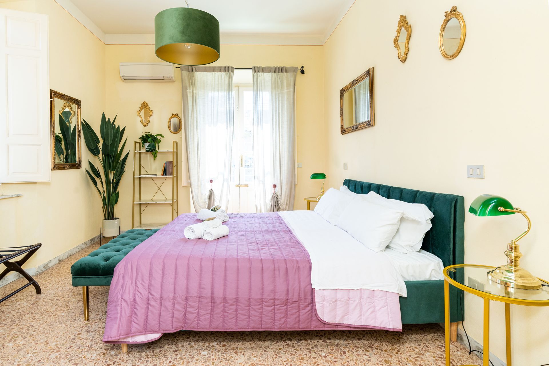 Bedroom with teal bed, gold accents, pink bedspread, green lamp, and large window.