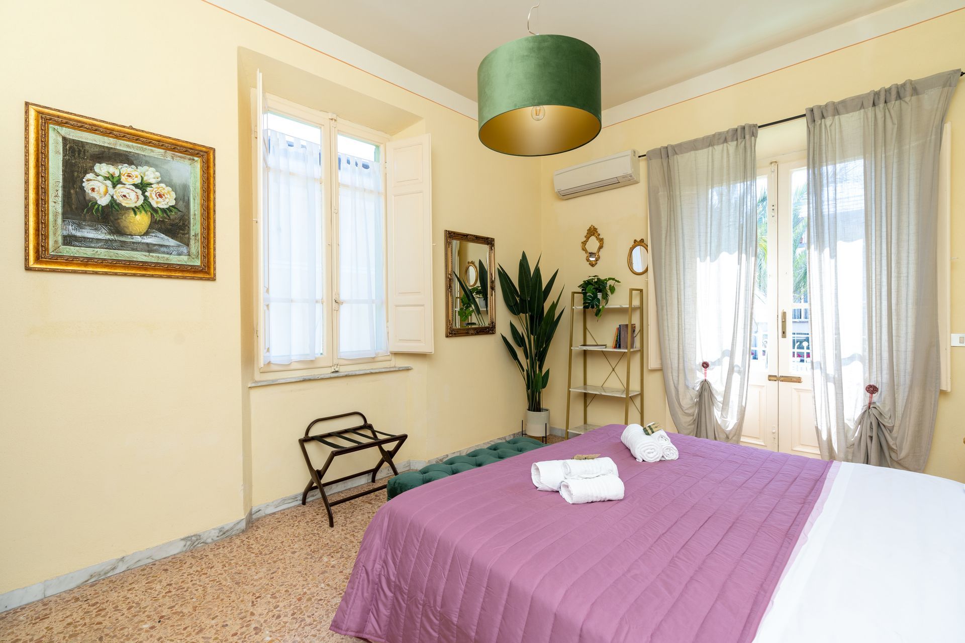 Bedroom with bed, art, windows, and decorative plants. Light walls and flooring.