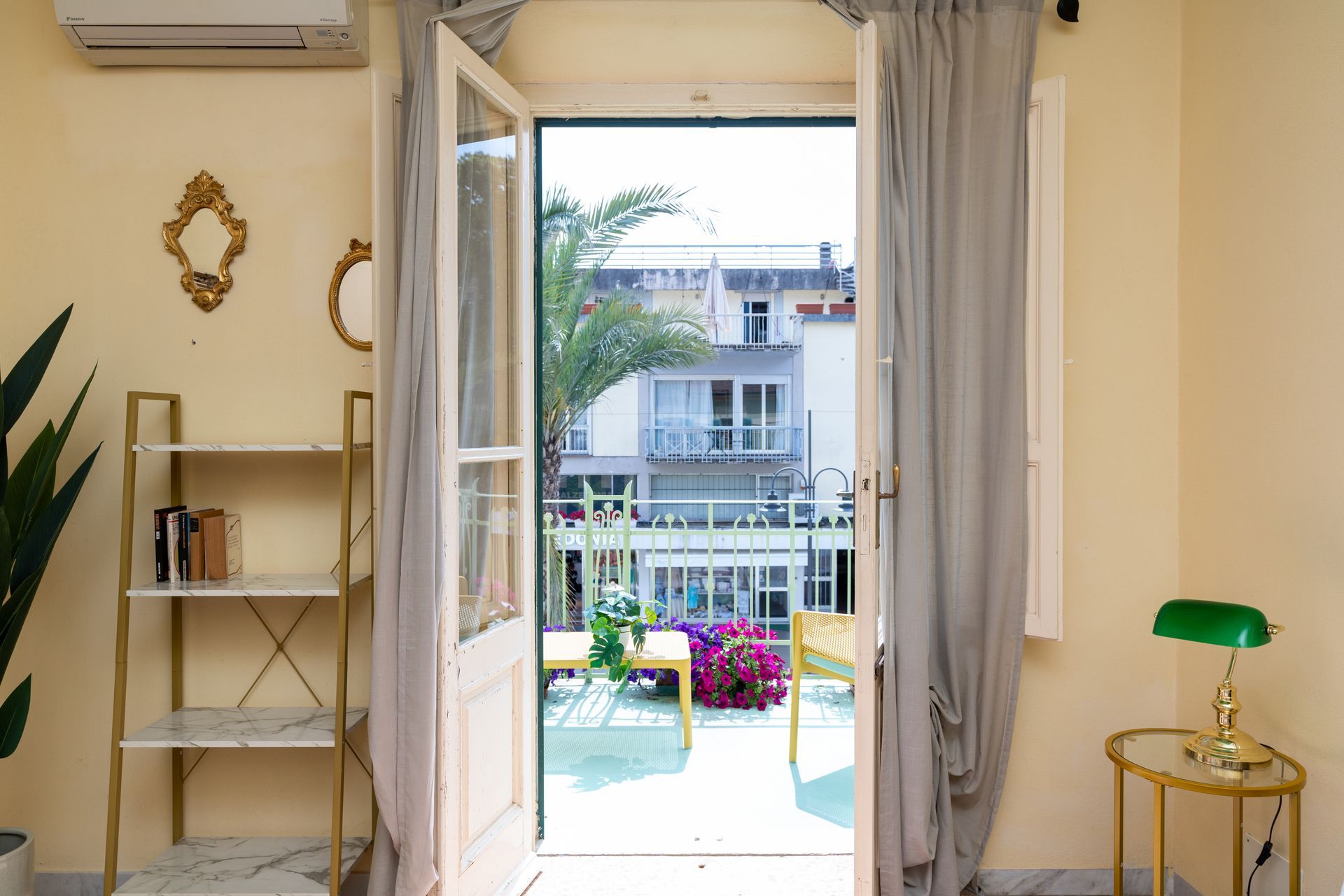 Open door to a balcony with white railing, plants, and small table. Interior has gold shelf and lamp.