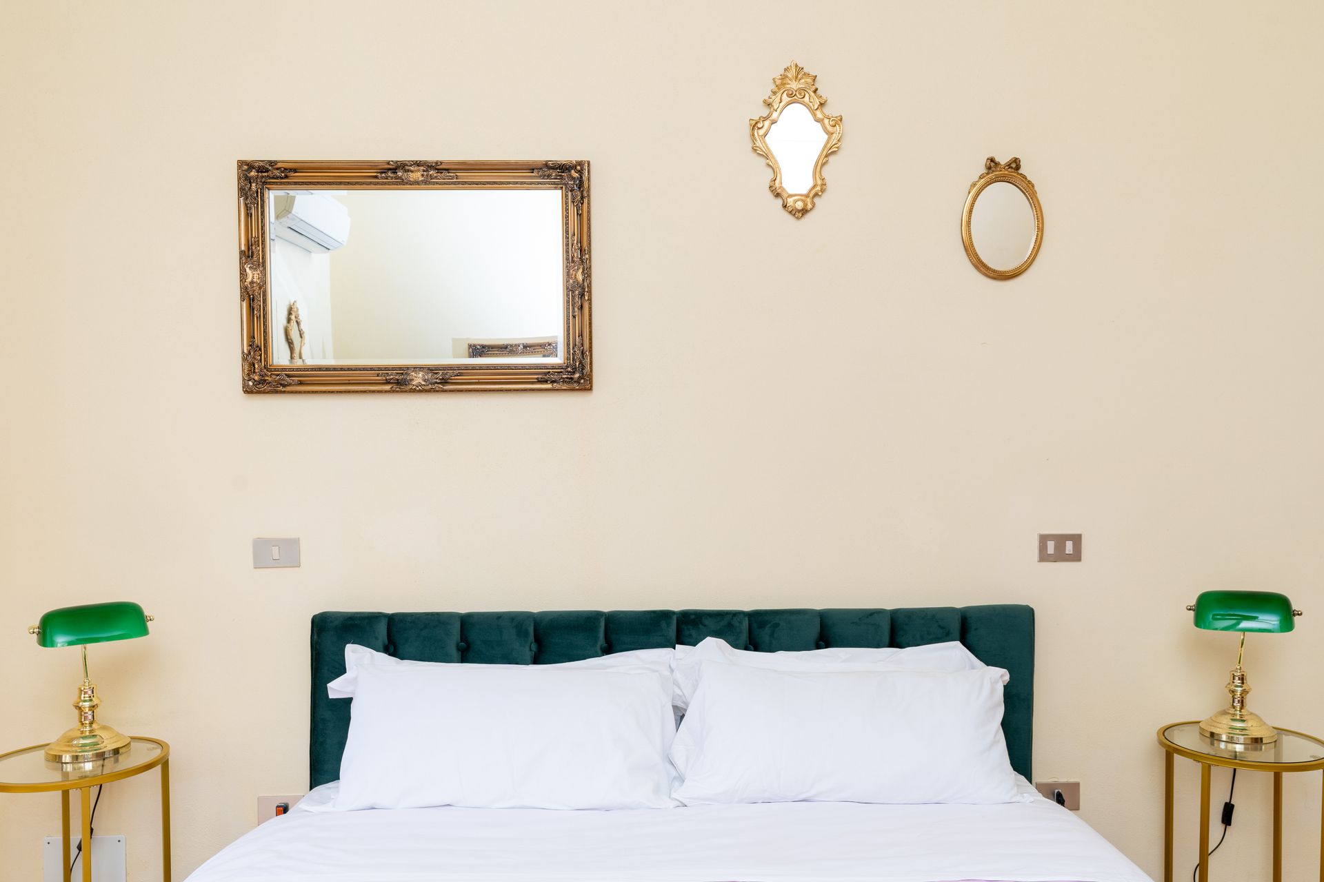 Bedroom with green tufted headboard, white bedding, and gold-framed mirrors and lamps.
