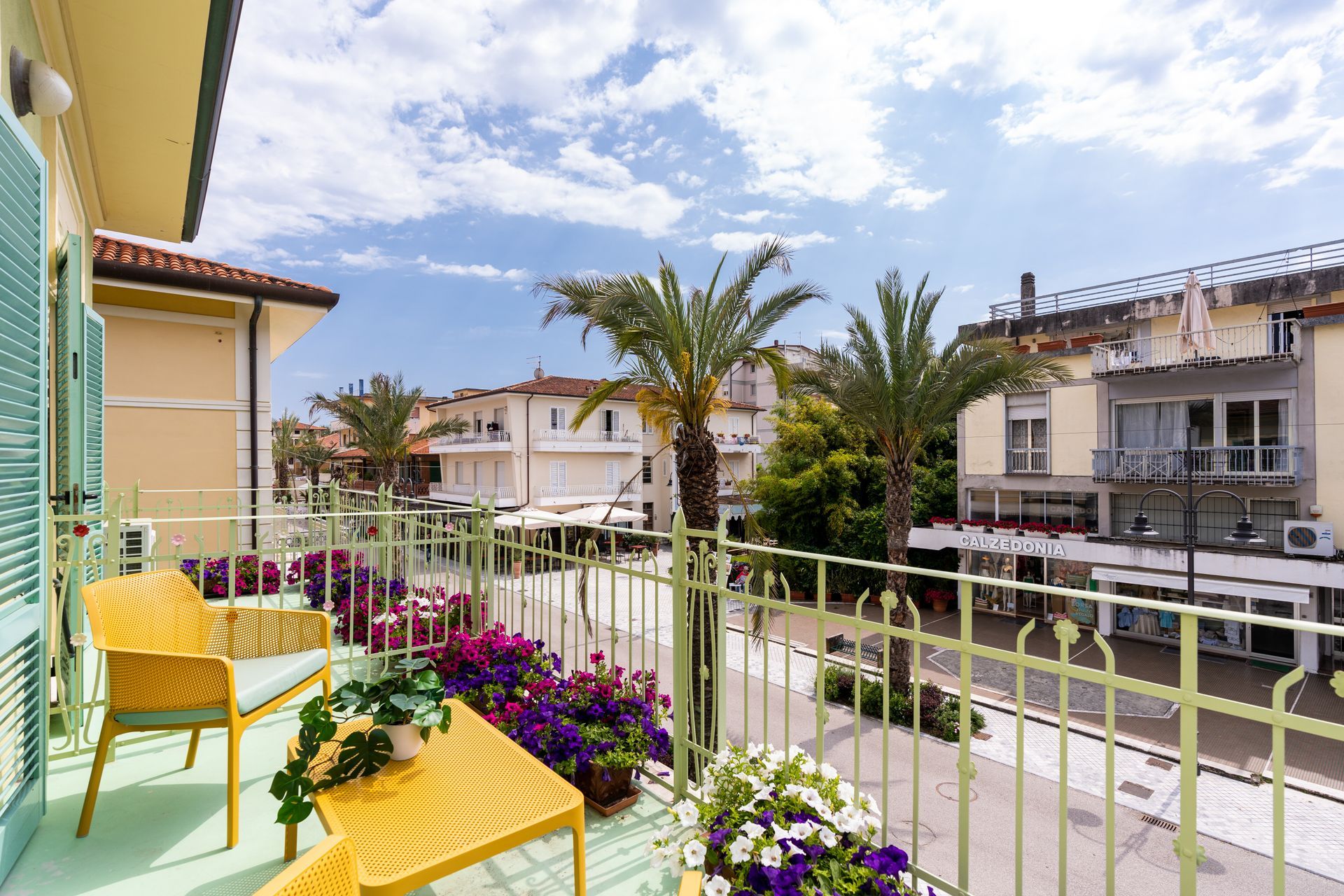 Balcony with yellow furniture, overlooking a street with buildings, palm trees, and flowers.