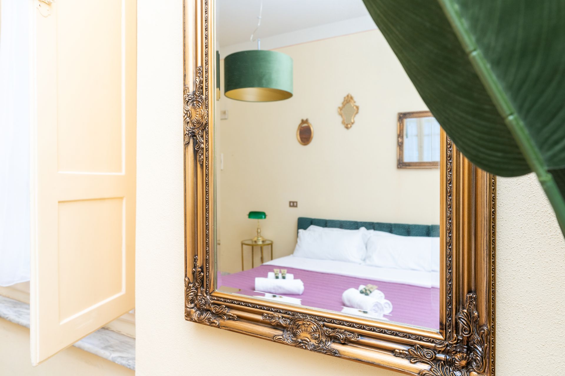 Mirror reflecting a bedroom with a bed, lamps, and decorative frames. A partially open doorway is on the left.