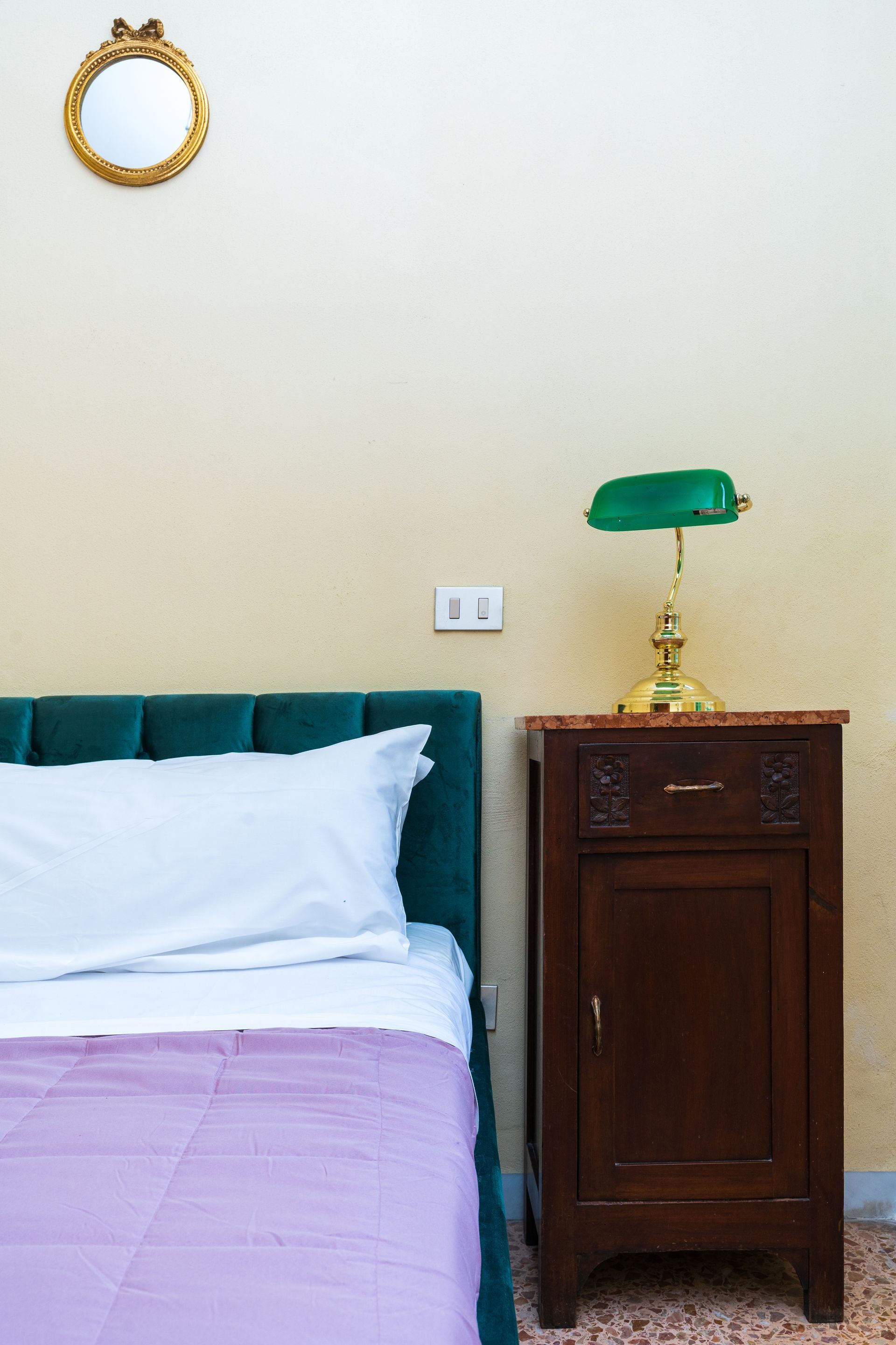 Bed with purple blanket, green headboard, and bedside table with green lamp and decorative mirror.