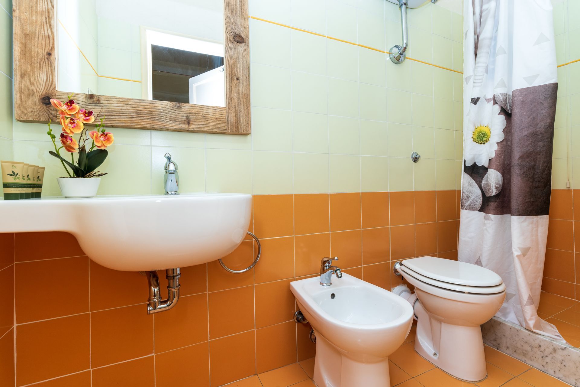 Bathroom with orange and green tiled walls, white sink, toilet, bidet, and shower curtain with flowers.