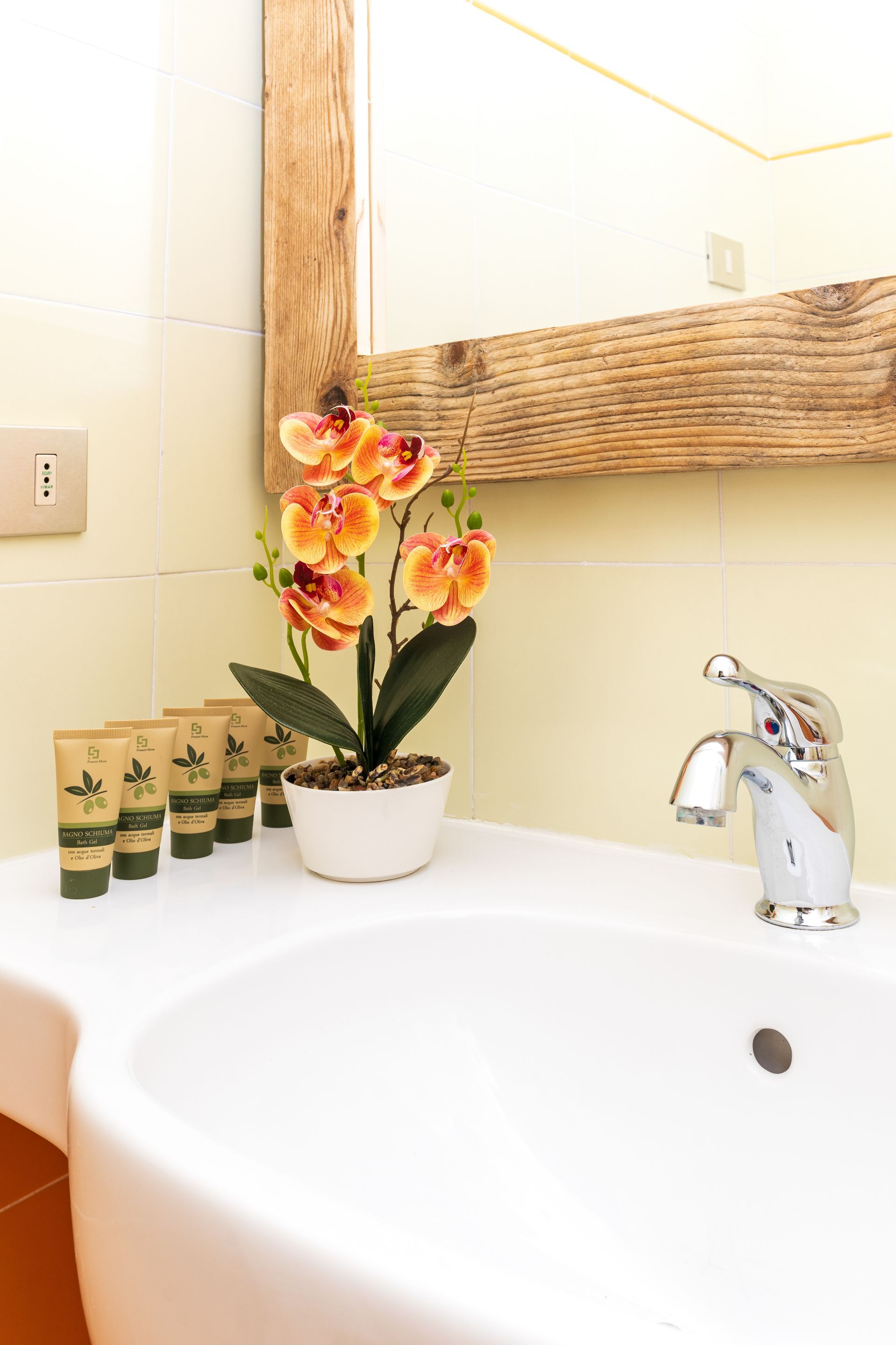White bathroom sink with faucet, toiletries, and an orange flower arrangement. A mirror is above the sink.