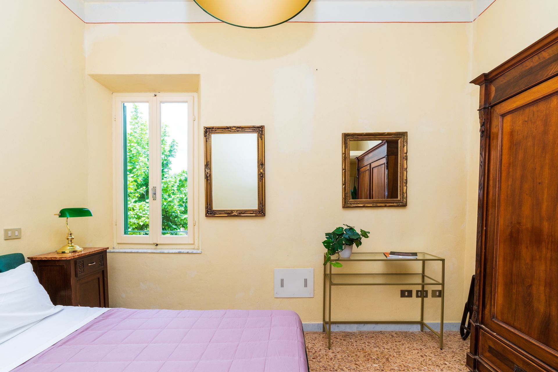 Bedroom with bed, antique furniture, mirrors, and window overlooking trees.