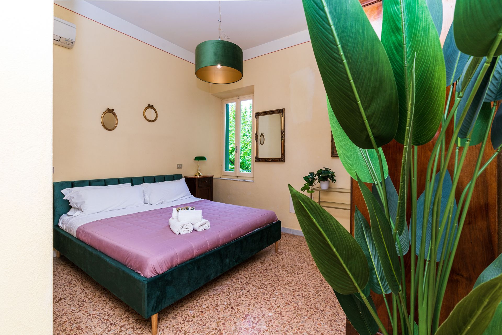 Bedroom with a green velvet bed, pink bedding, a green lampshade, and a large indoor plant.