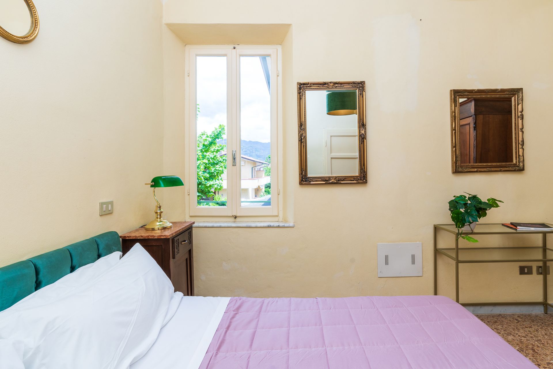 Bedroom with bed, mirrors, window, and small table. Pink bedspread, teal headboard, cream walls.