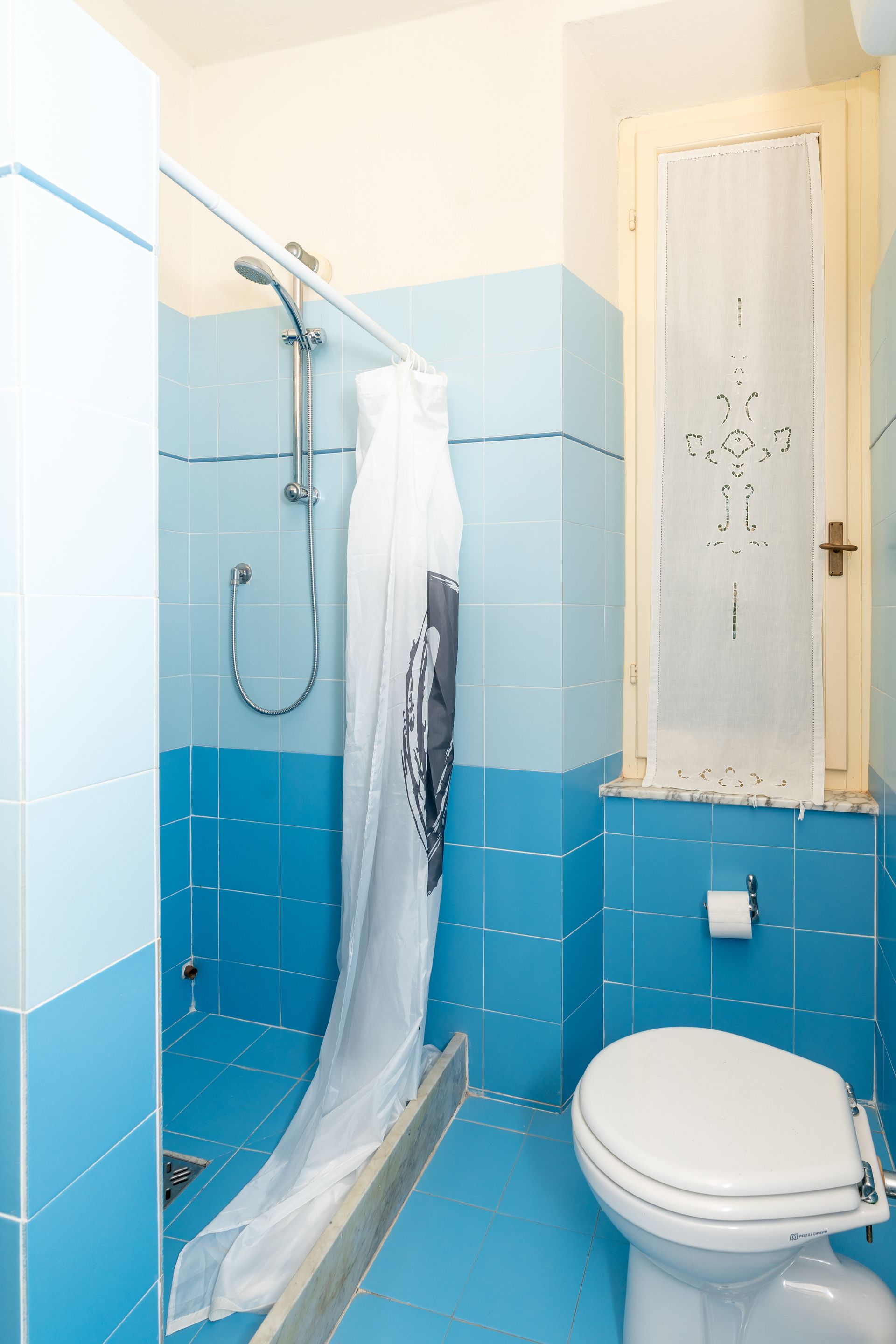 Blue-tiled bathroom with shower, toilet, and window with a white curtain.