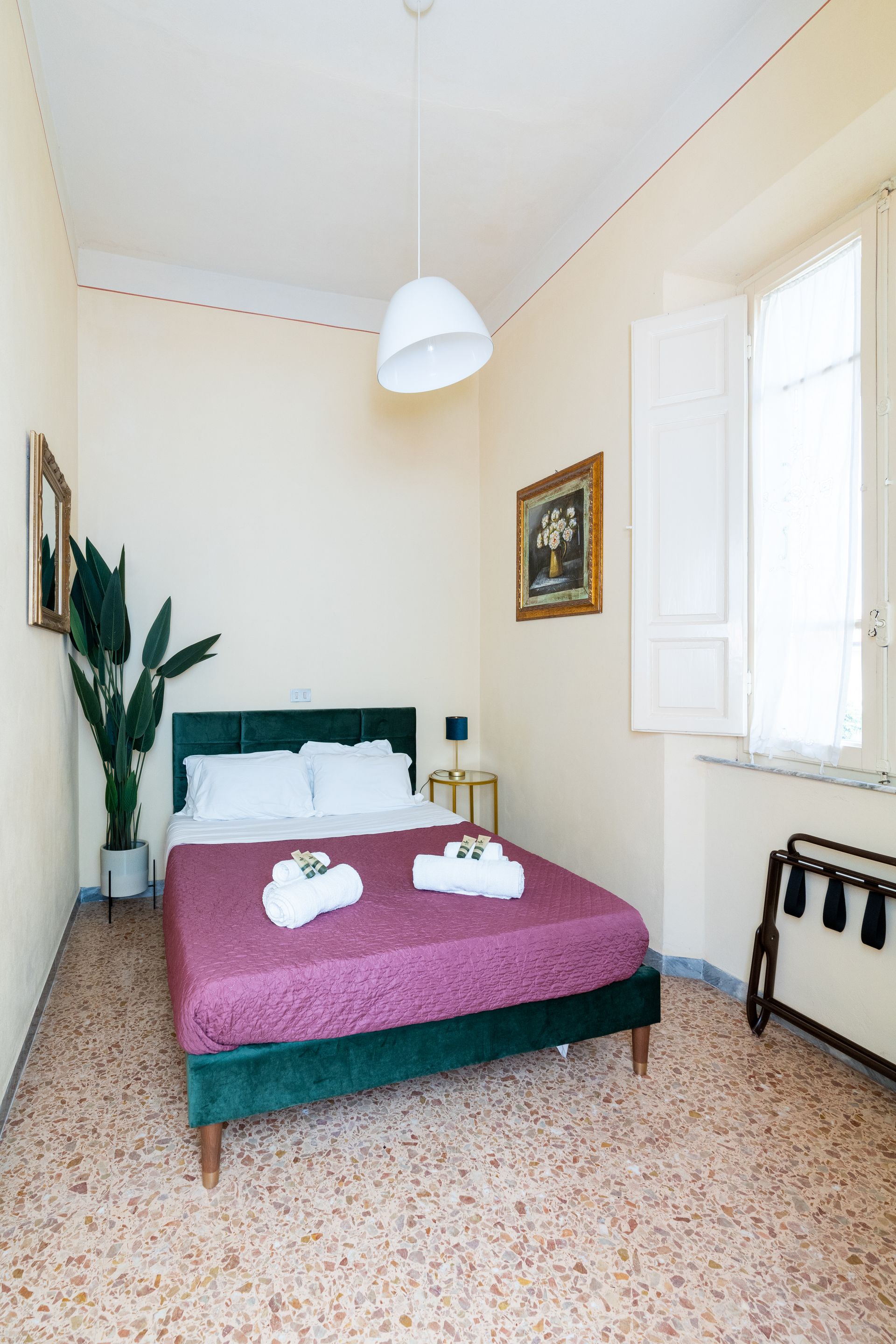 Bedroom with a green bed, purple blanket, white towels, a window with shutters, and a tall plant.