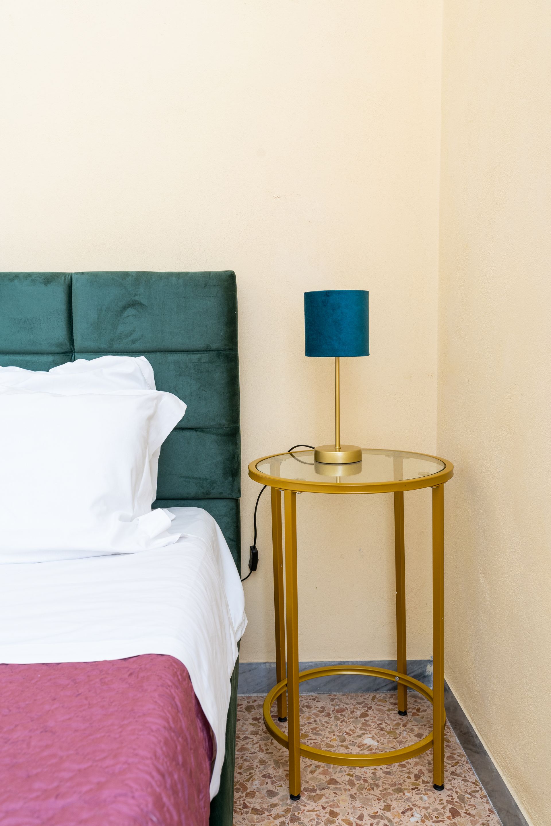 Bed with green headboard, white bedding, and pink blanket. Gold side table with blue lamp.