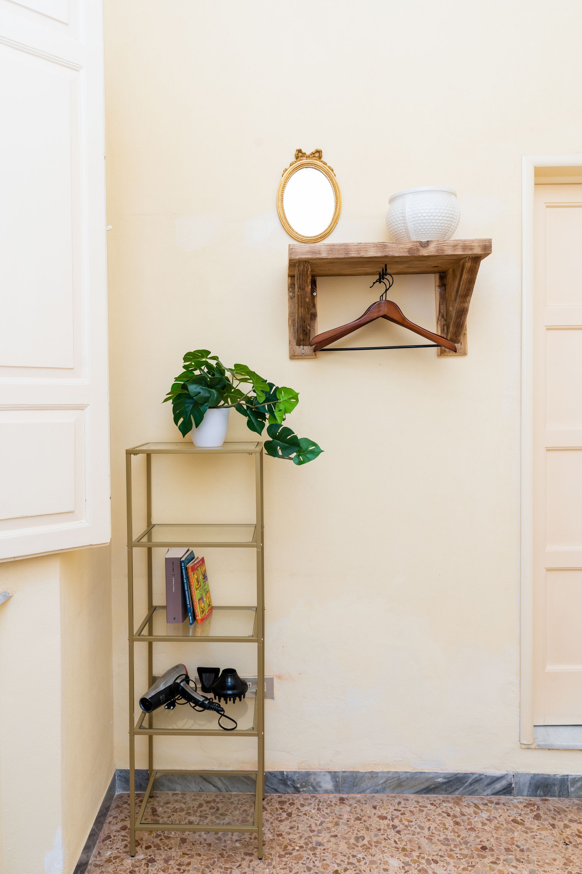 A gold metal shelf with decor, a wooden shelf with a mirror, and a potted plant against a yellow wall.