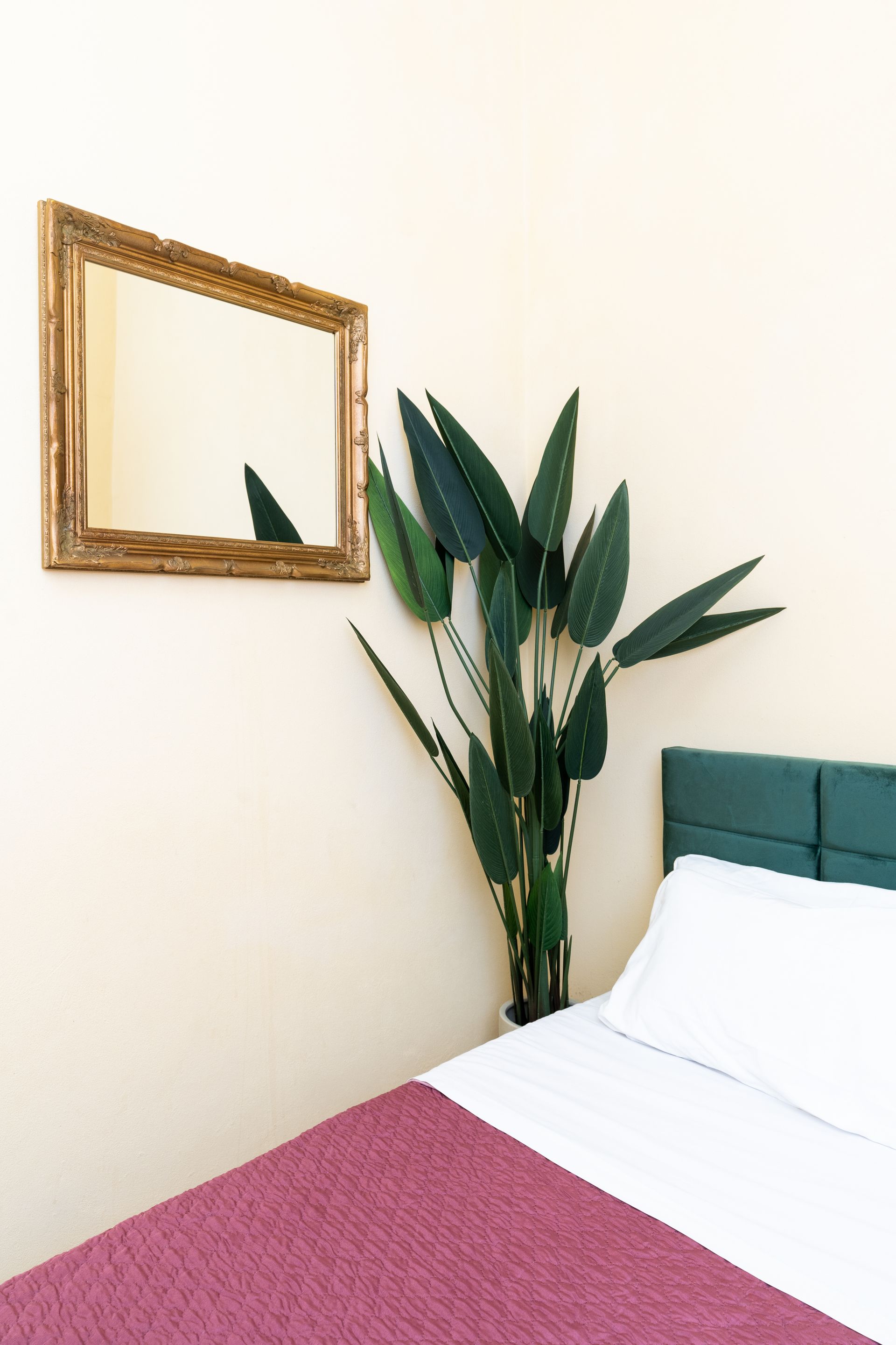 Bedroom corner with gold-framed mirror, tall green plant, and bed with white linens and red blanket.