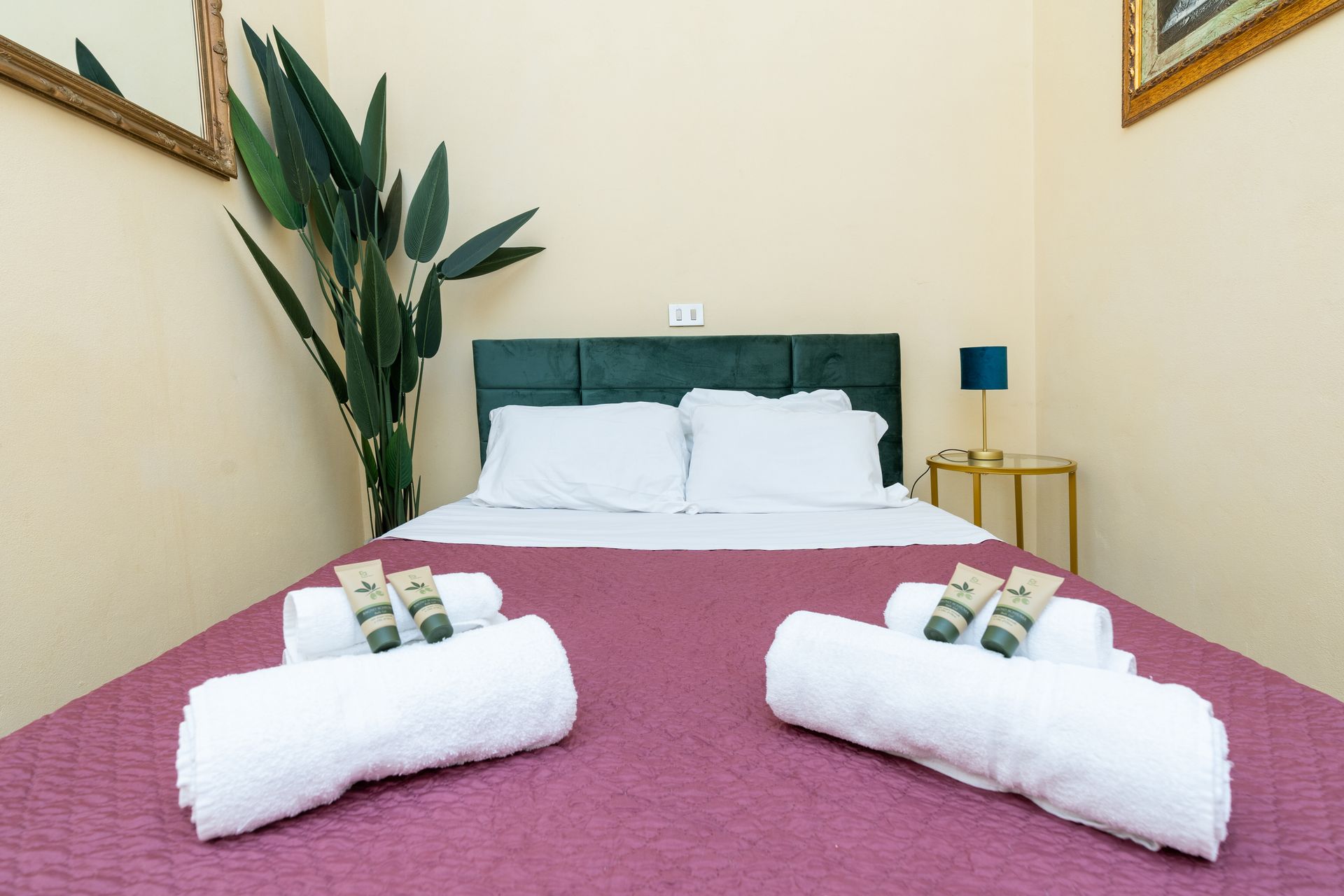 Bedroom with bed, towels, green plant, and small gold side table with lamp.