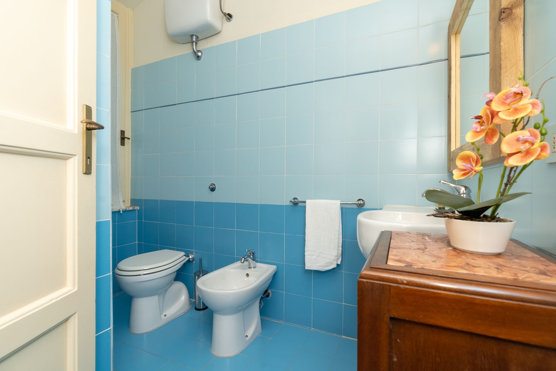 Bathroom with blue tile walls, a toilet, bidet, sink, mirror, and decorative flowers.