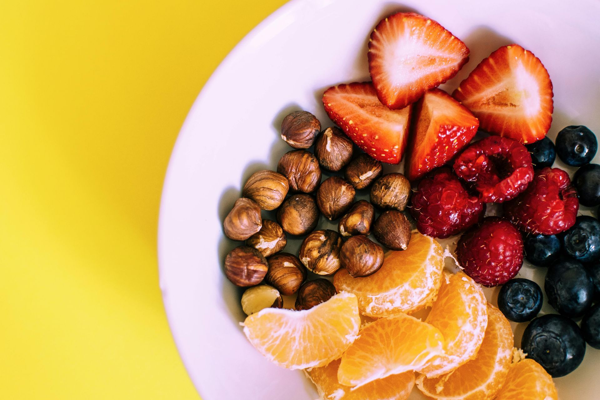Plate with strawberries, raspberries, blueberries, hazelnuts, and tangerine slices on a yellow surface.