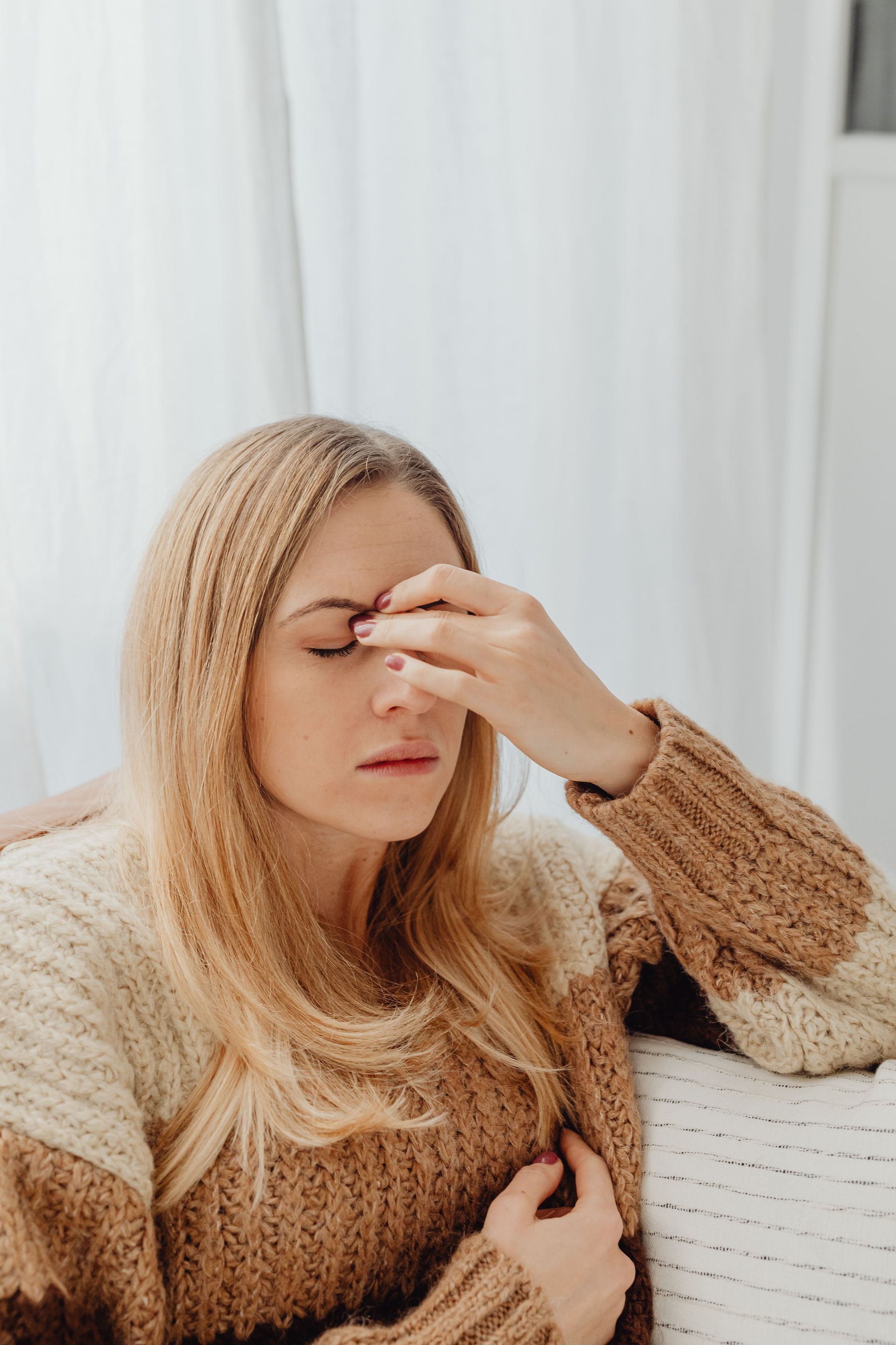 Woman holding her forehead on a white sofa, looking tired in a cozy beige sweater
