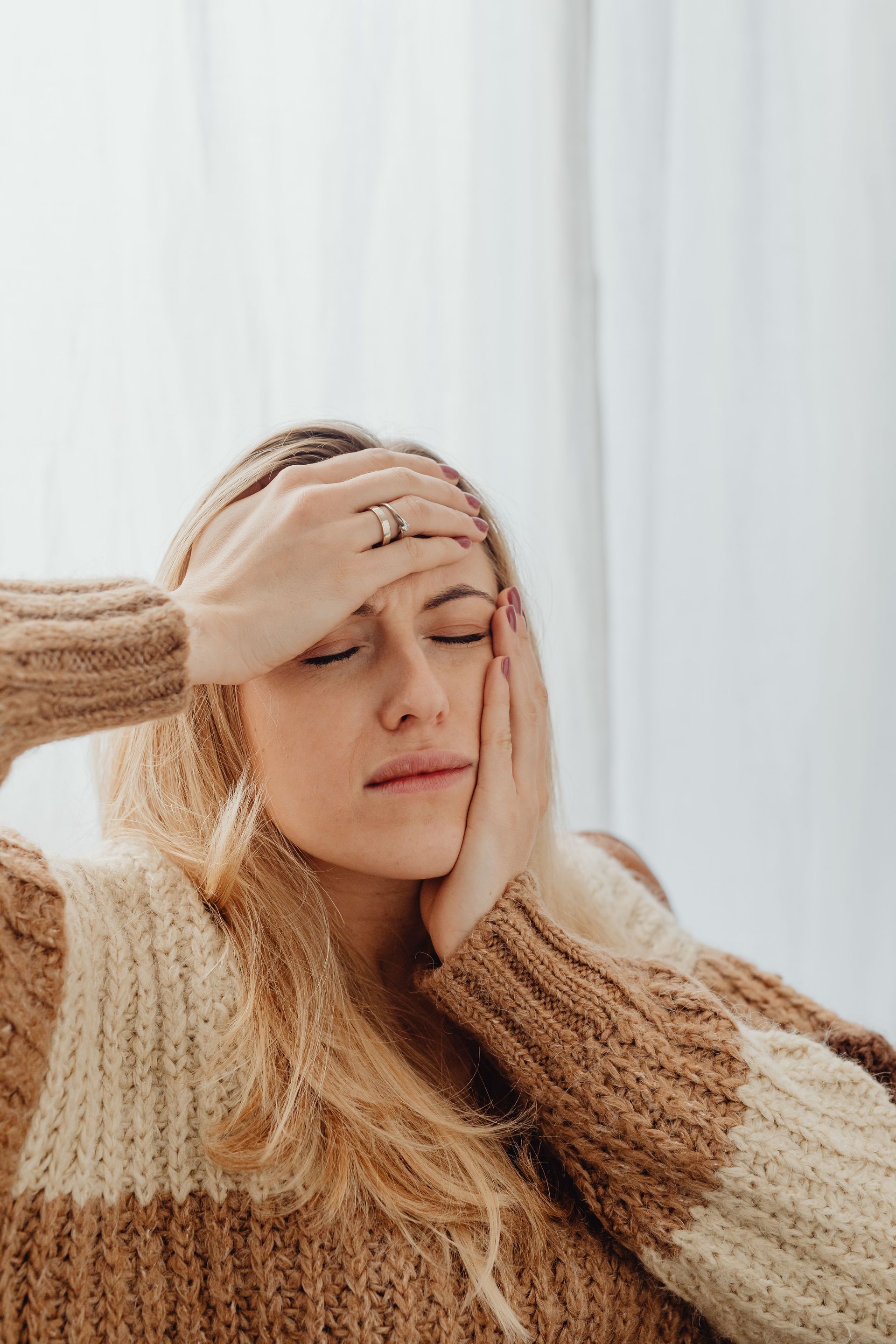 Person in a beige sweater holding their forehead and face, looking stressed against a light curtain background