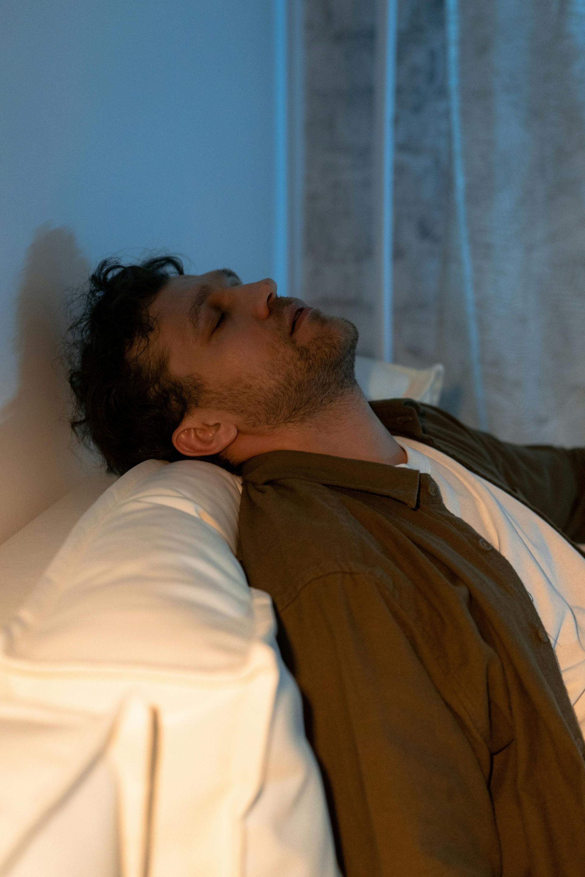 Man reclines on a pillow by a curtained window, eyes closed, in a dimly lit room.