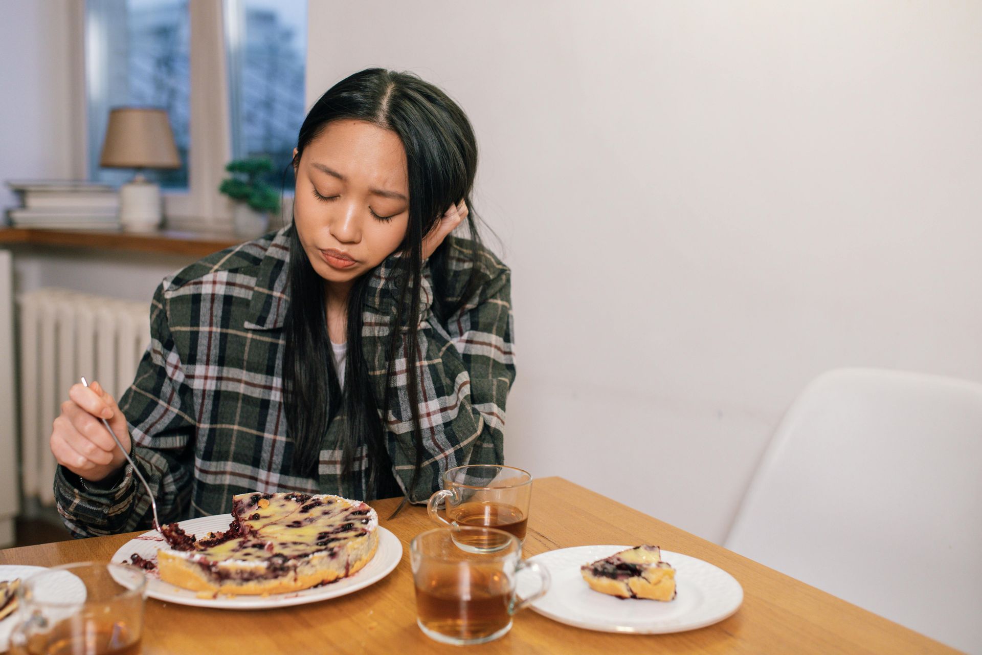 Woman in plaid eating pie, looking thoughtful at a table, tea nearby.