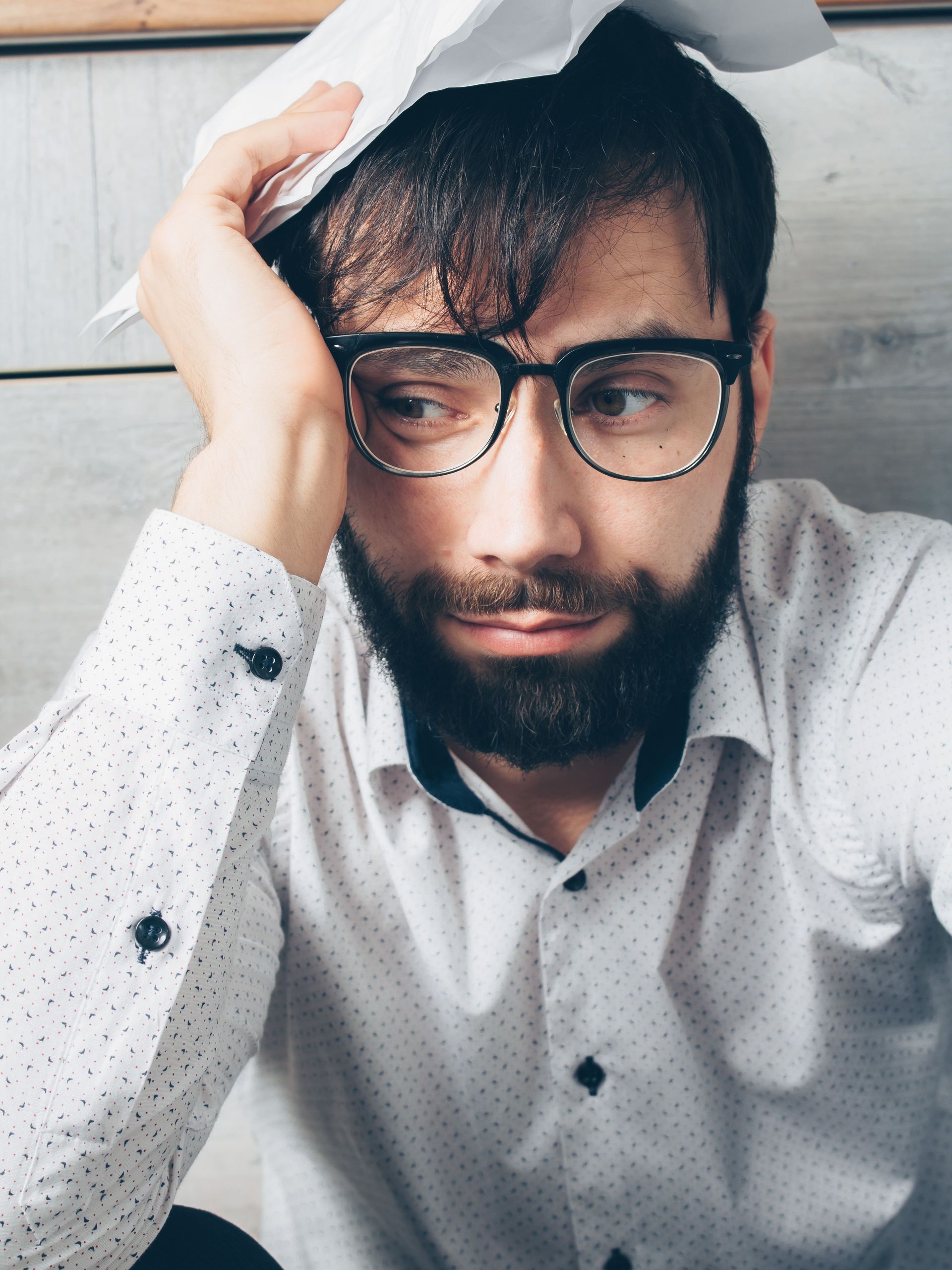 Tired man with glasses holding his head under a white cloth, looking down in a close-up portrait.